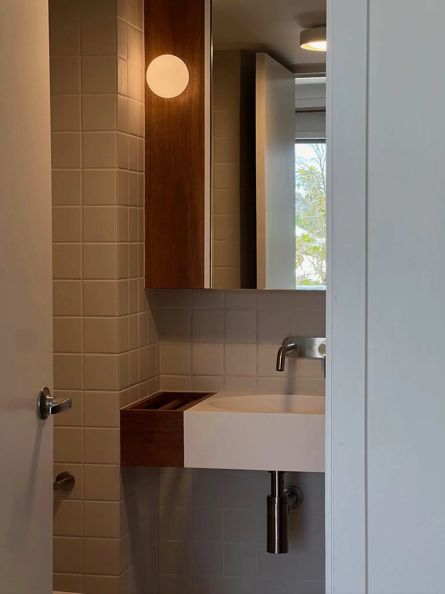 Small bathroom with a white sink, a mirror, a wooden cabinet, beige tiled walls, and a window showing trees outside.