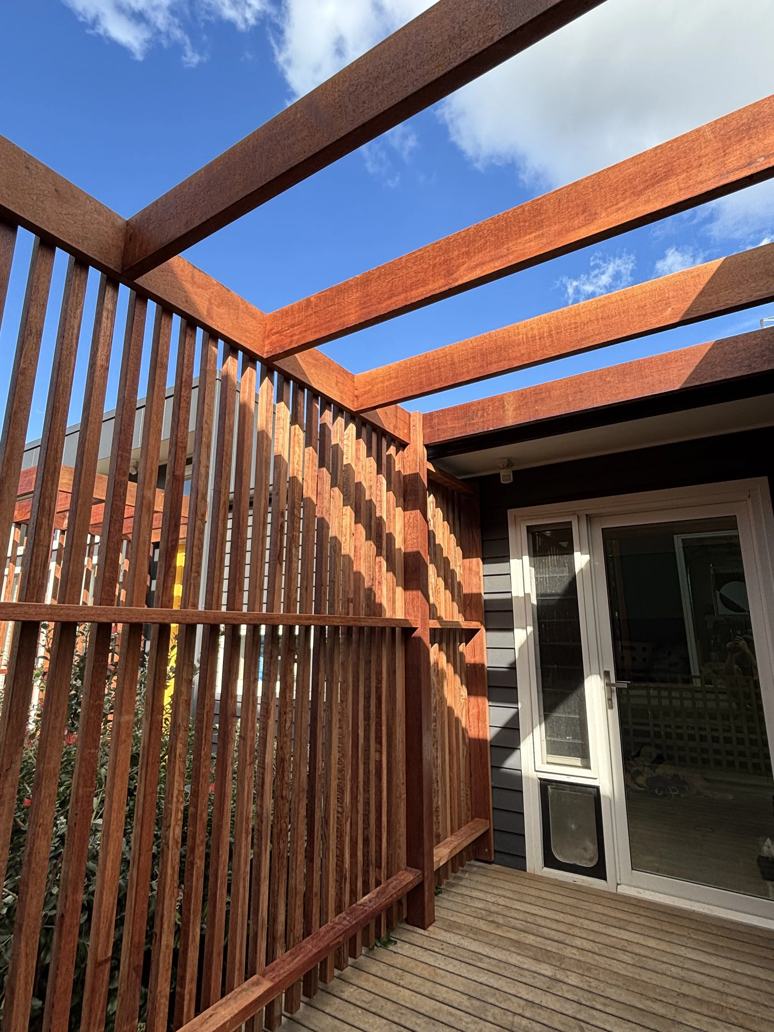 Wooden deck with vertical slats and horizontal beams, connected to house with sliding glass door, under a blue sky with some clouds.