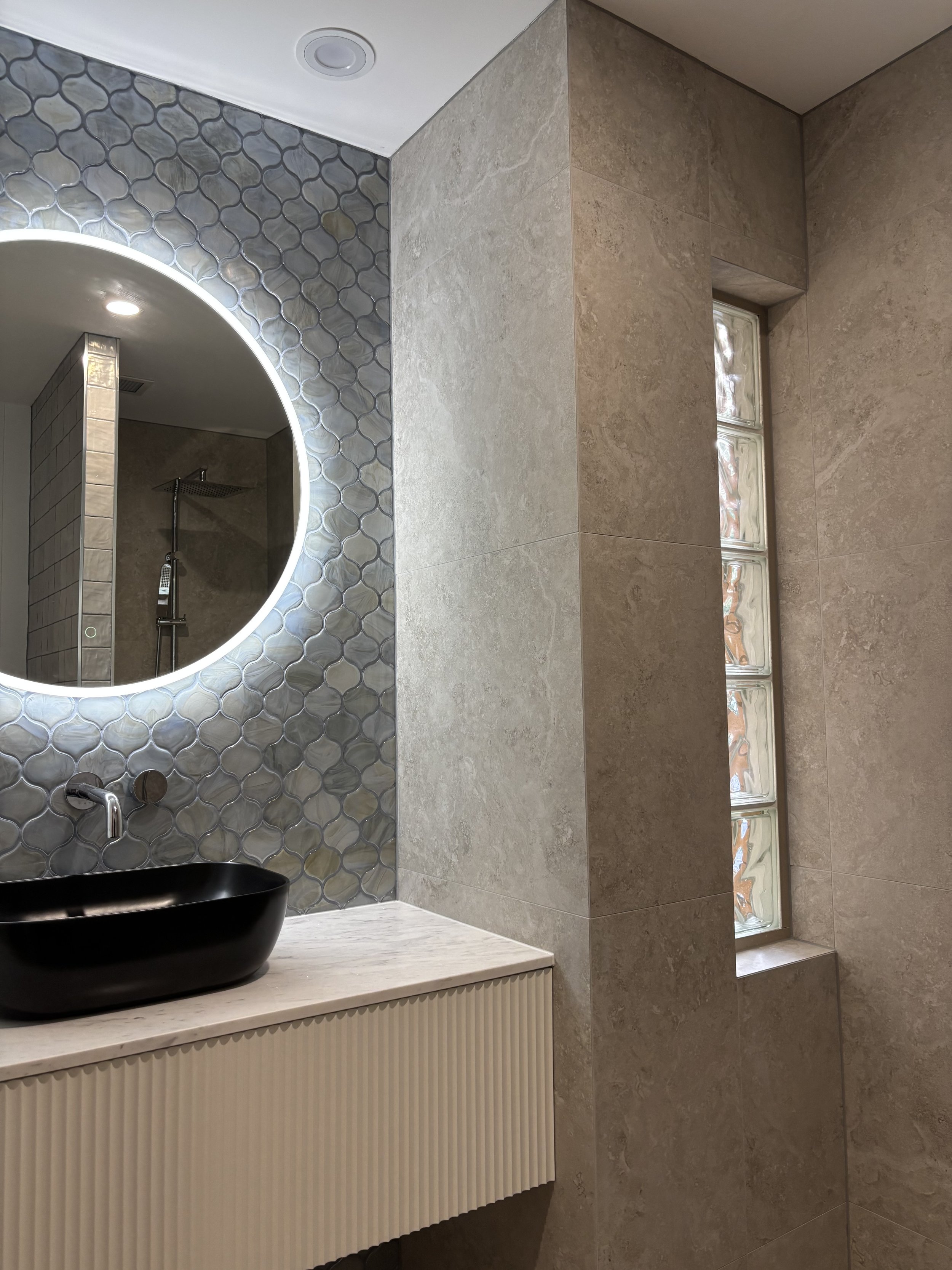 Modern bathroom with beige tiles, a circular mirror with backlight, a black vessel sink on a white marble countertop, a wall-mounted faucet, a glass block window, and a shower area with a rainfall showerhead.
