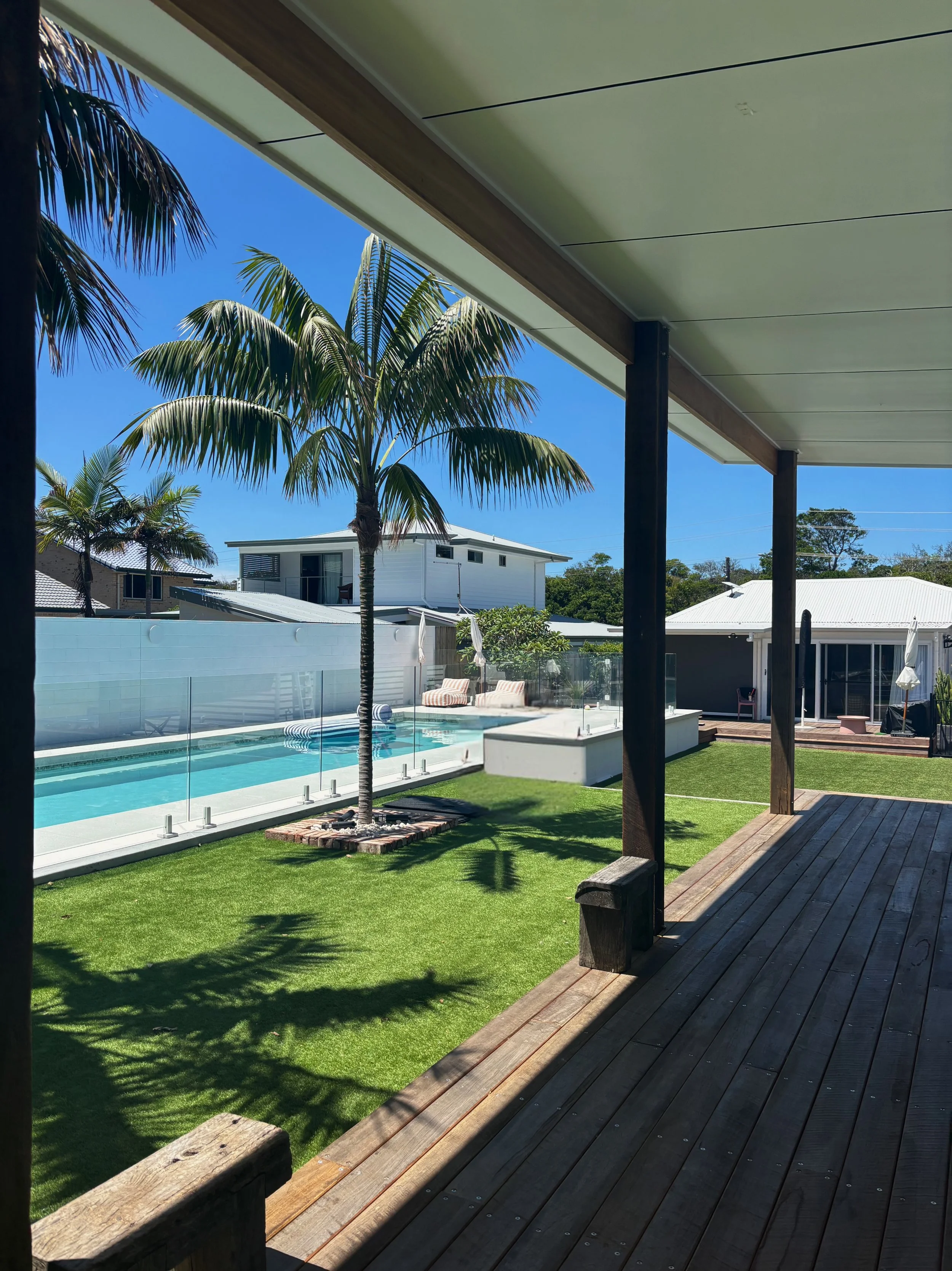 View of a backyard with a swimming pool, palm trees, green grass, and outdoor furniture, taken from a shaded porch.
