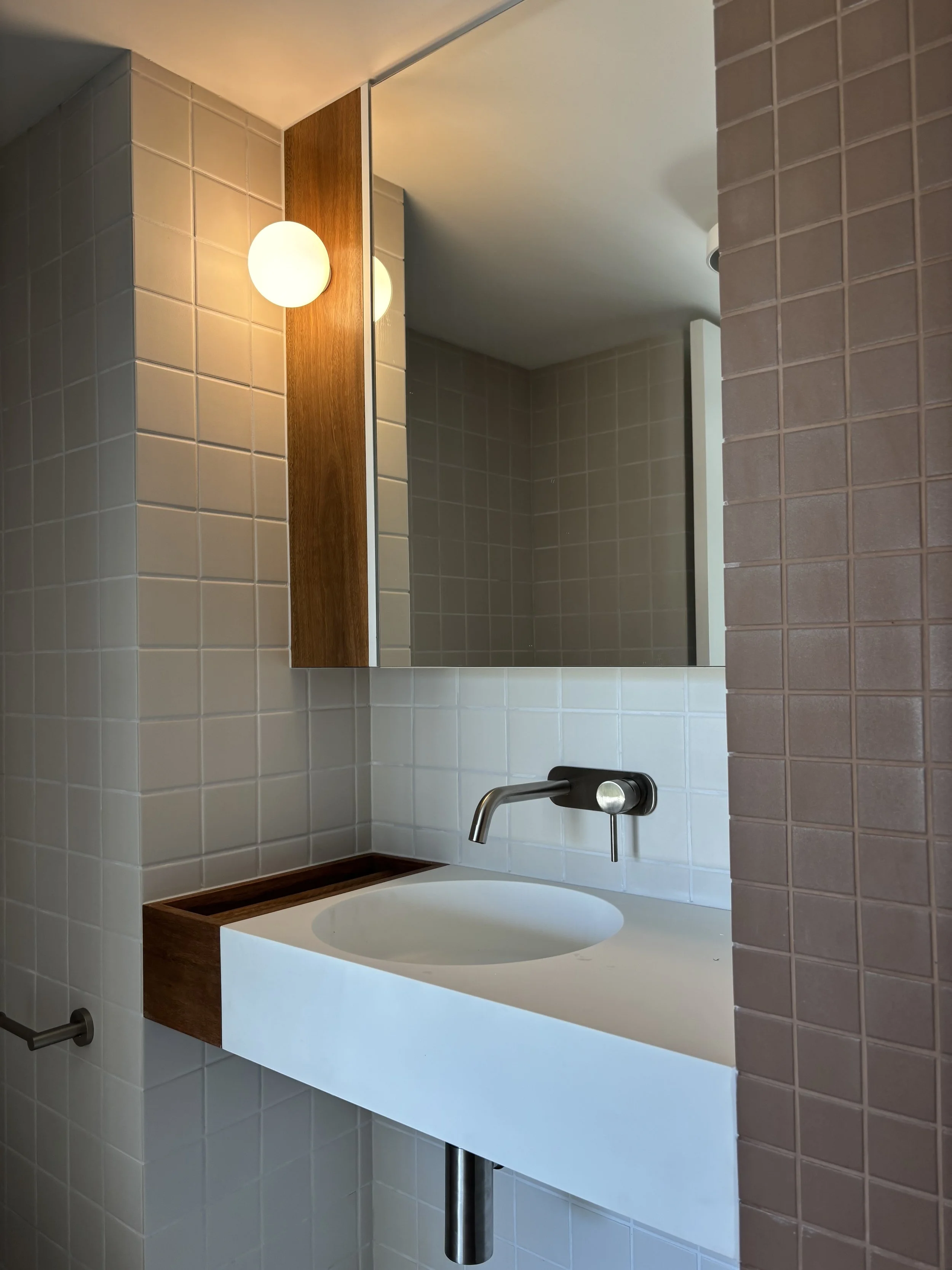 Bathroom with white rectangular sink, wall-mounted faucet, square beige tiles, a large mirror, and a wooden accent with spherical wall light.