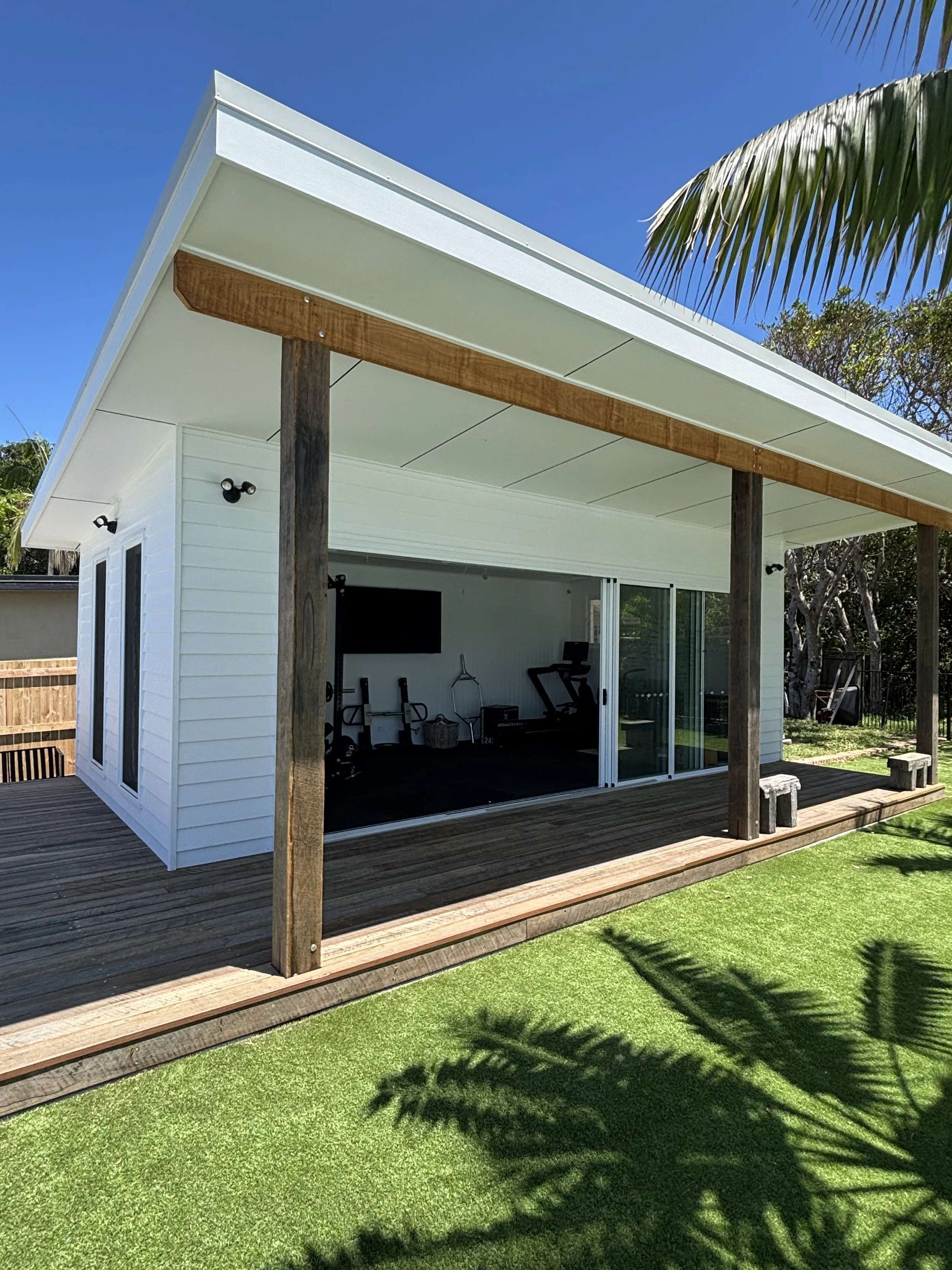 Backyard patio with a wooden deck, white house with large sliding glass doors, exercise equipment inside, and a shadow of a palm tree on green grass.