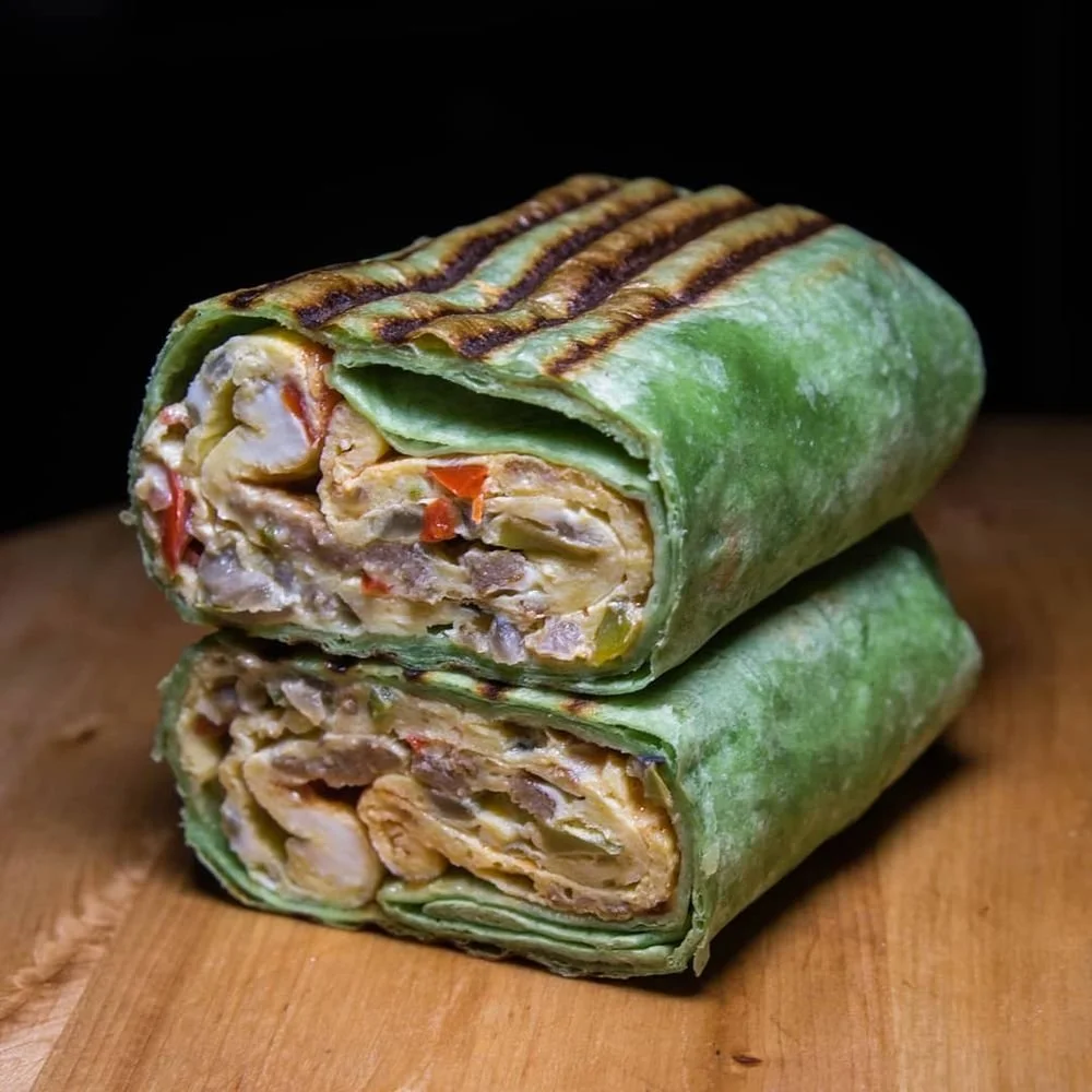 Two grilled wrap sandwiches with a green flour tortilla, filled with chicken, vegetables, and sauce, sliced in half, on a wooden surface against a black background.