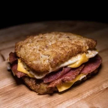 A breakfast sandwich with fried chicken, bacon, scrambled eggs, and melted cheese on a bread bun.