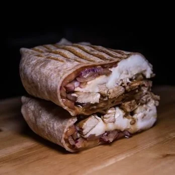 A close-up of a sliced wrap sandwich with grilled bread, bacon, melted cheese, and possibly turkey or chicken, on a wooden surface.