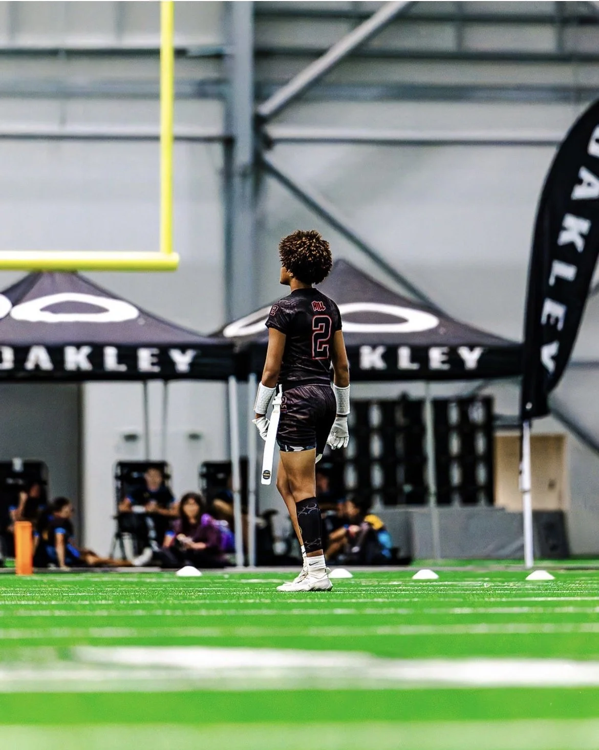 A female football player in black uniform standing on a green indoor football field, facing away from the camera, with a prosthetic leg, surrounded by tents and spectators.