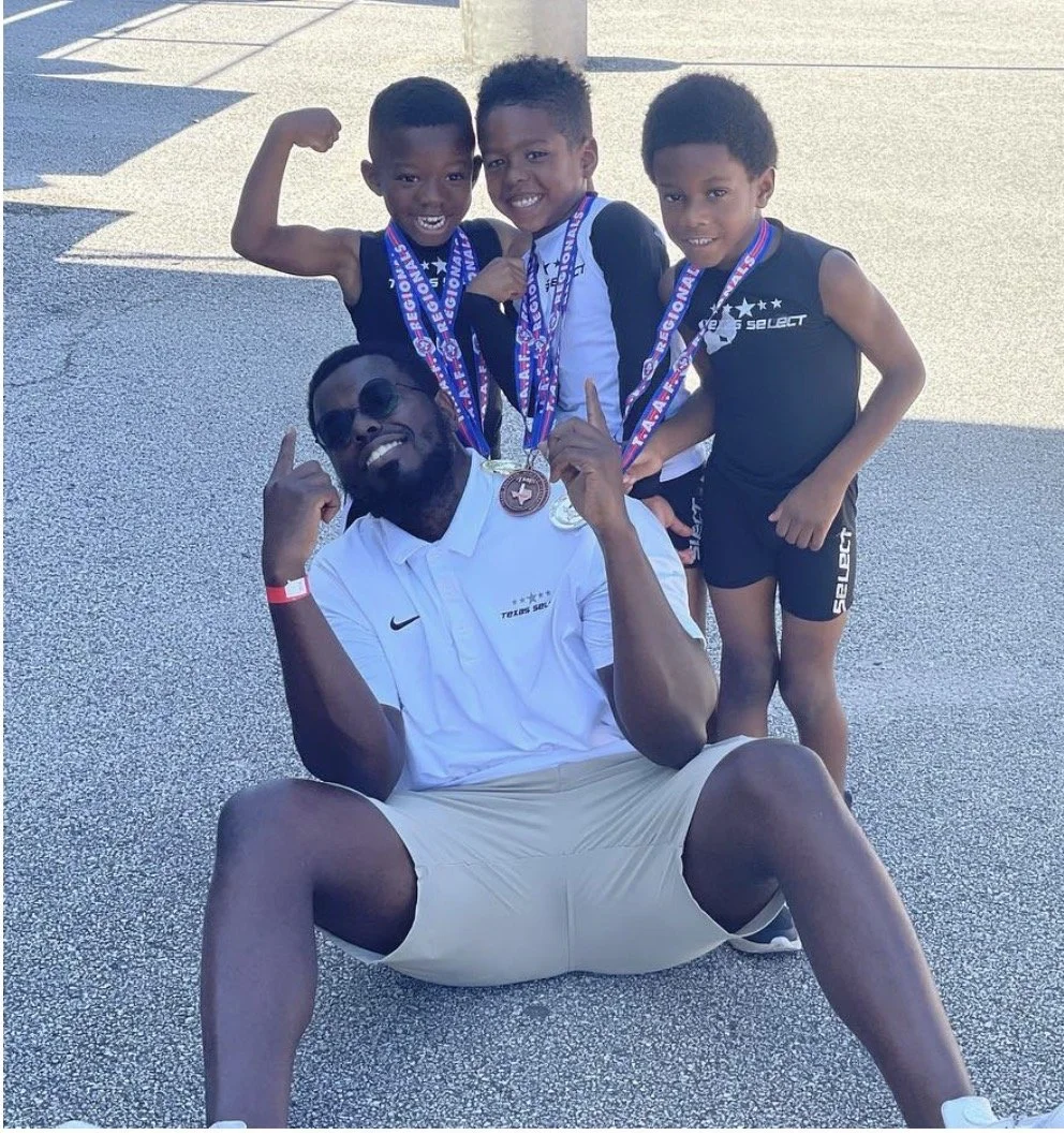 Four young boys and one adult man are posing outdoors, with the man sitting on the ground and flexing his arm. All of them are smiling and wearing medals around their necks, indicating they participated in a sporting event. The boys are in athletic clothing, and the man is wearing sunglasses and a white polo shirt.