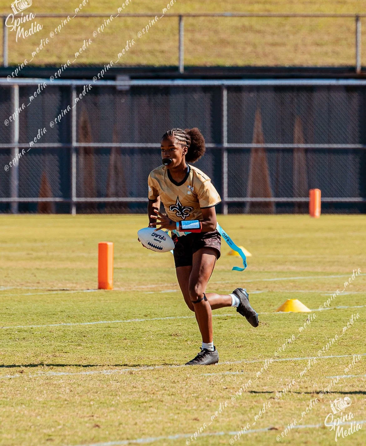 A young girl running on a football field during a training session with a football in her hands, wearing a gold and black sports jersey.