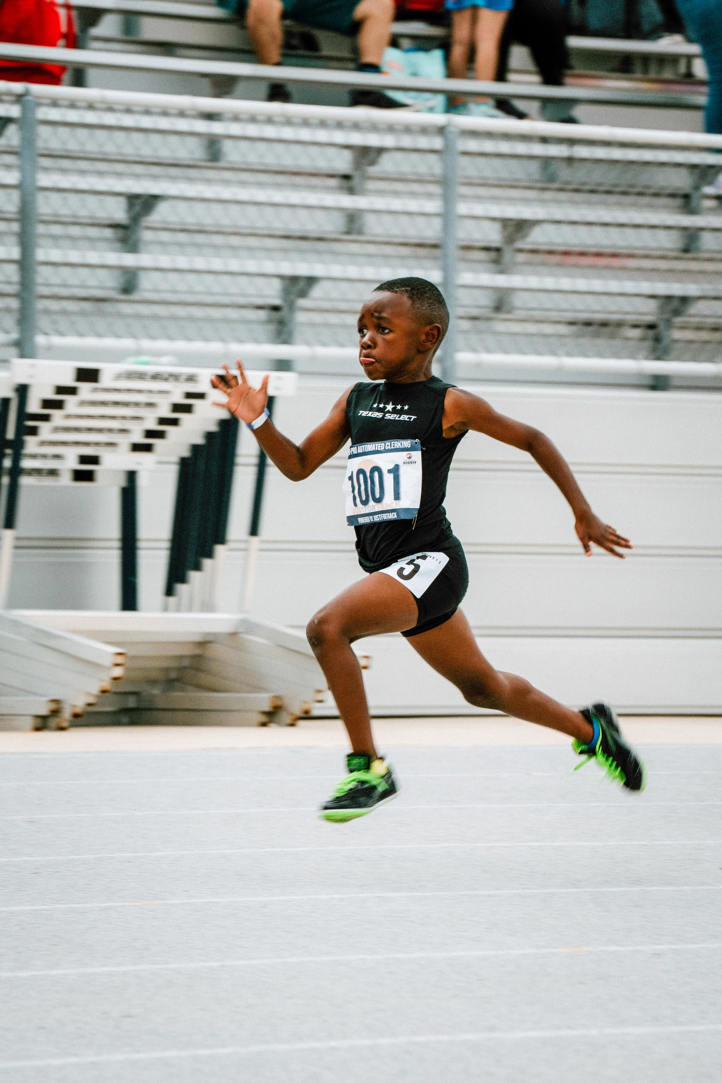 A young boy running in a race, wearing a black athletic uniform with the number 101, on a track with hurdles in the background and spectators sitting on bleachers.