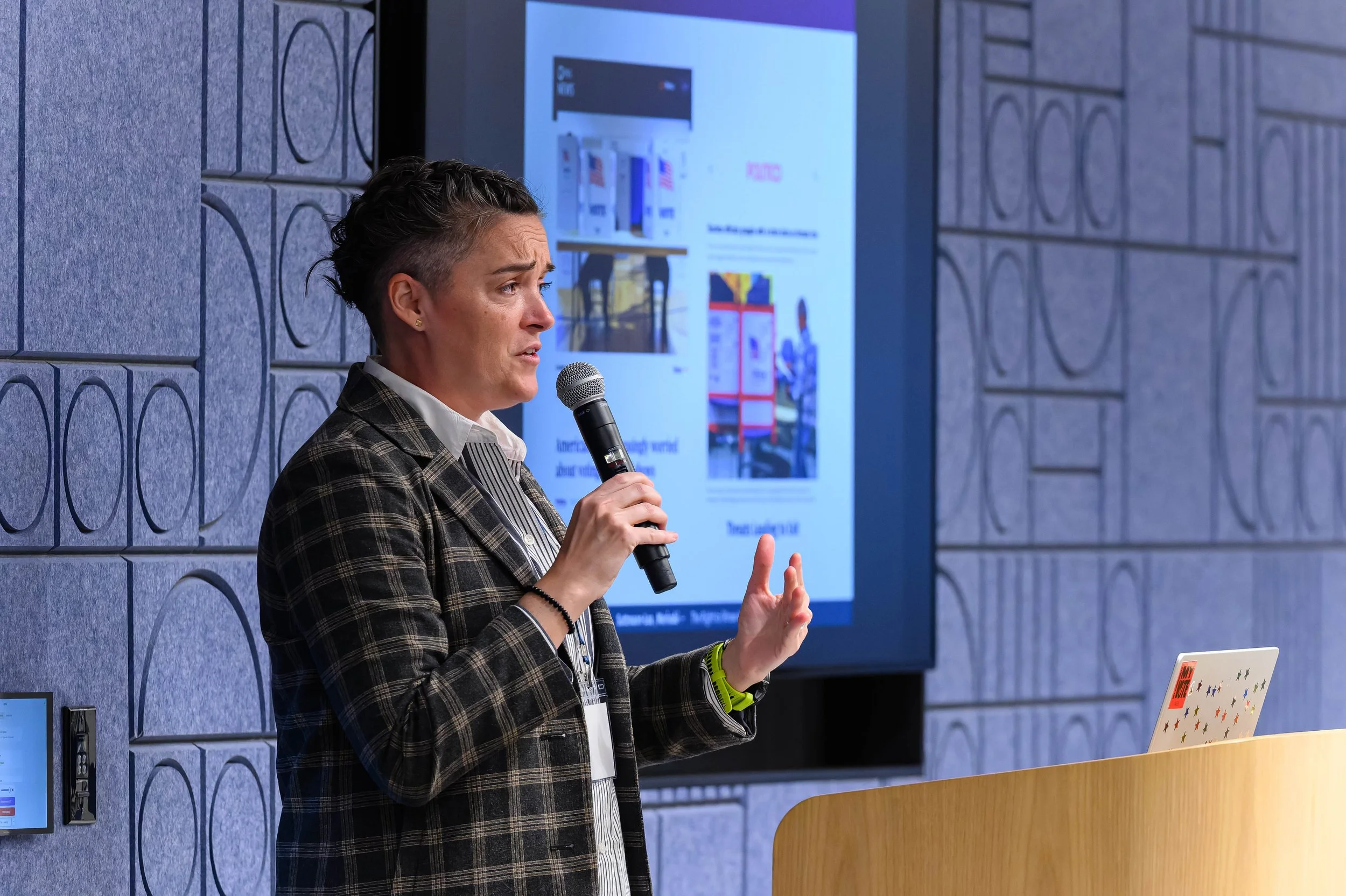 A woman with short dark hair presenting with a microphone in front of a large screen, wearing a plaid blazer and a white shirt, in a conference room with textured blue walls.