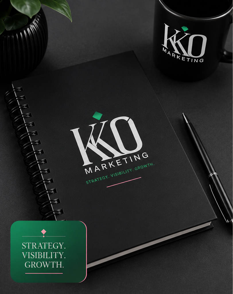 A black spiral notebook with the words 'KKO MARKETING' and the subtitle 'STRATEGY. VISIBILITY. GROWTH.' on the cover, a black pen, a black coffee mug with the same logo, and a black vase with green leaves, on a dark table.