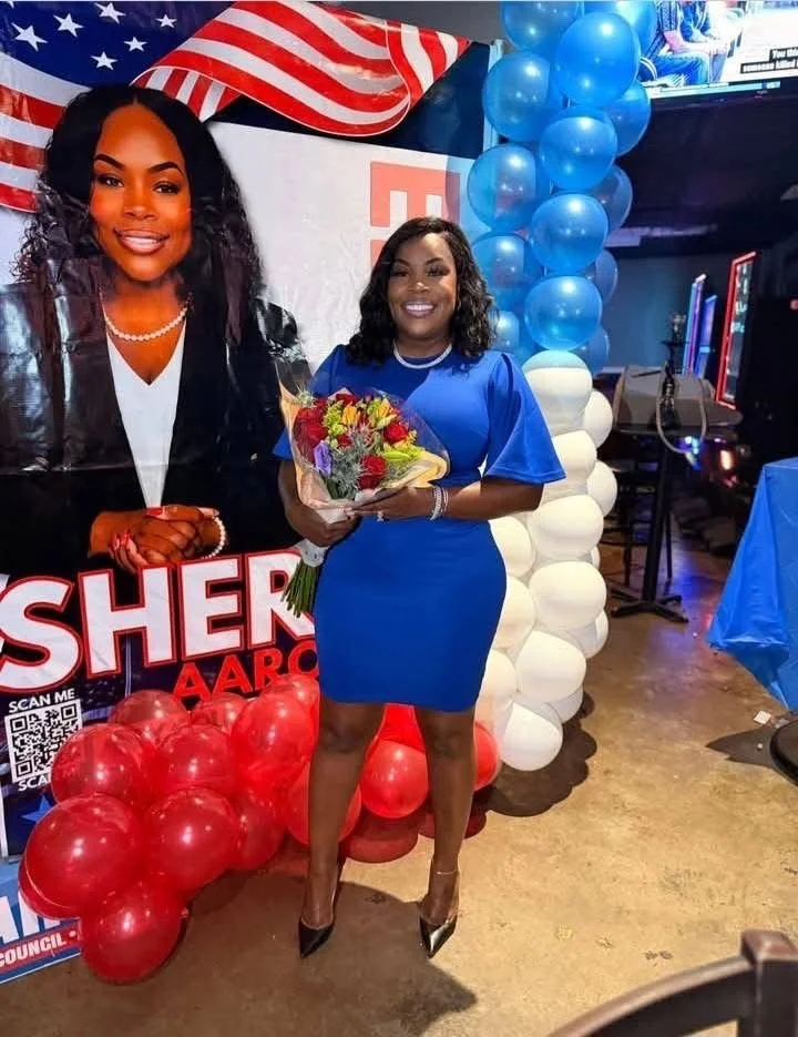 A woman in a royal blue dress with black heels holding a bouquet of flowers, standing in front of a political campaign display featuring a large poster of a woman and balloons in red, white, and blue.