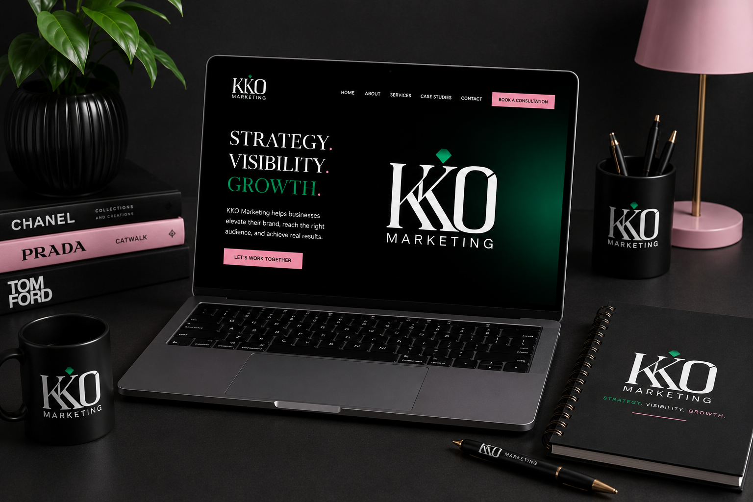 A workspace featuring a MacBook displaying the KKO Marketing website, with matching black mug, notebook, and pen all branded with the KKO logo, alongside a stack of fashion-related books, a pink desk lamp with gold accents, a black pen holder, and a potted plant.