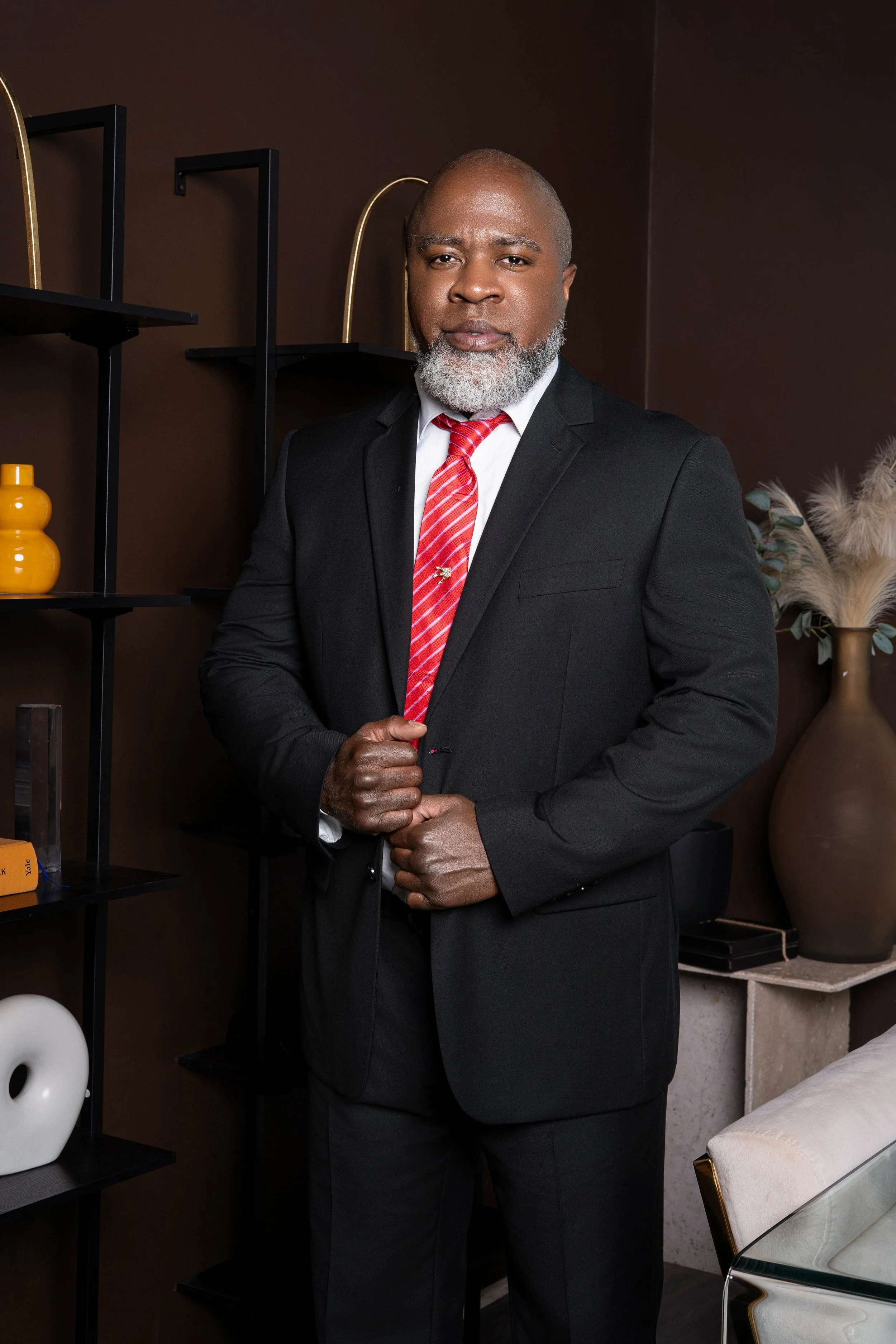 A confident Black man with a gray beard in a black suit and red striped tie, standing with hands clasped, in a modern interior with black shelves and decorative objects.