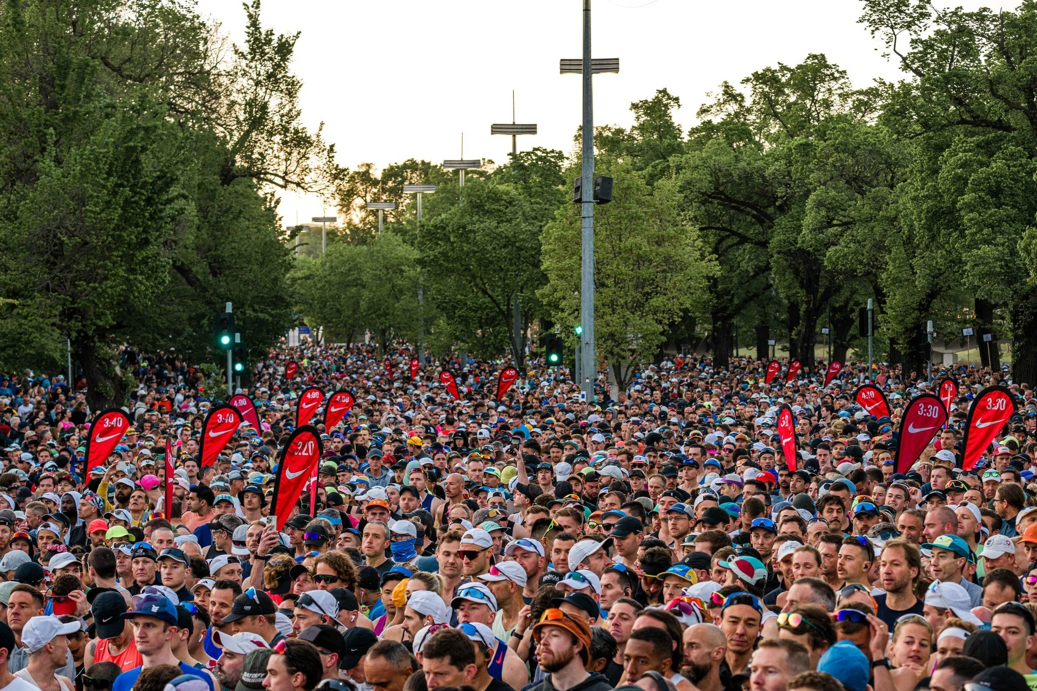 A large crowd of people gathered outdoors in a park, wearing athletic gear and hats, participating in a running event, with traffic lights and trees in the background.