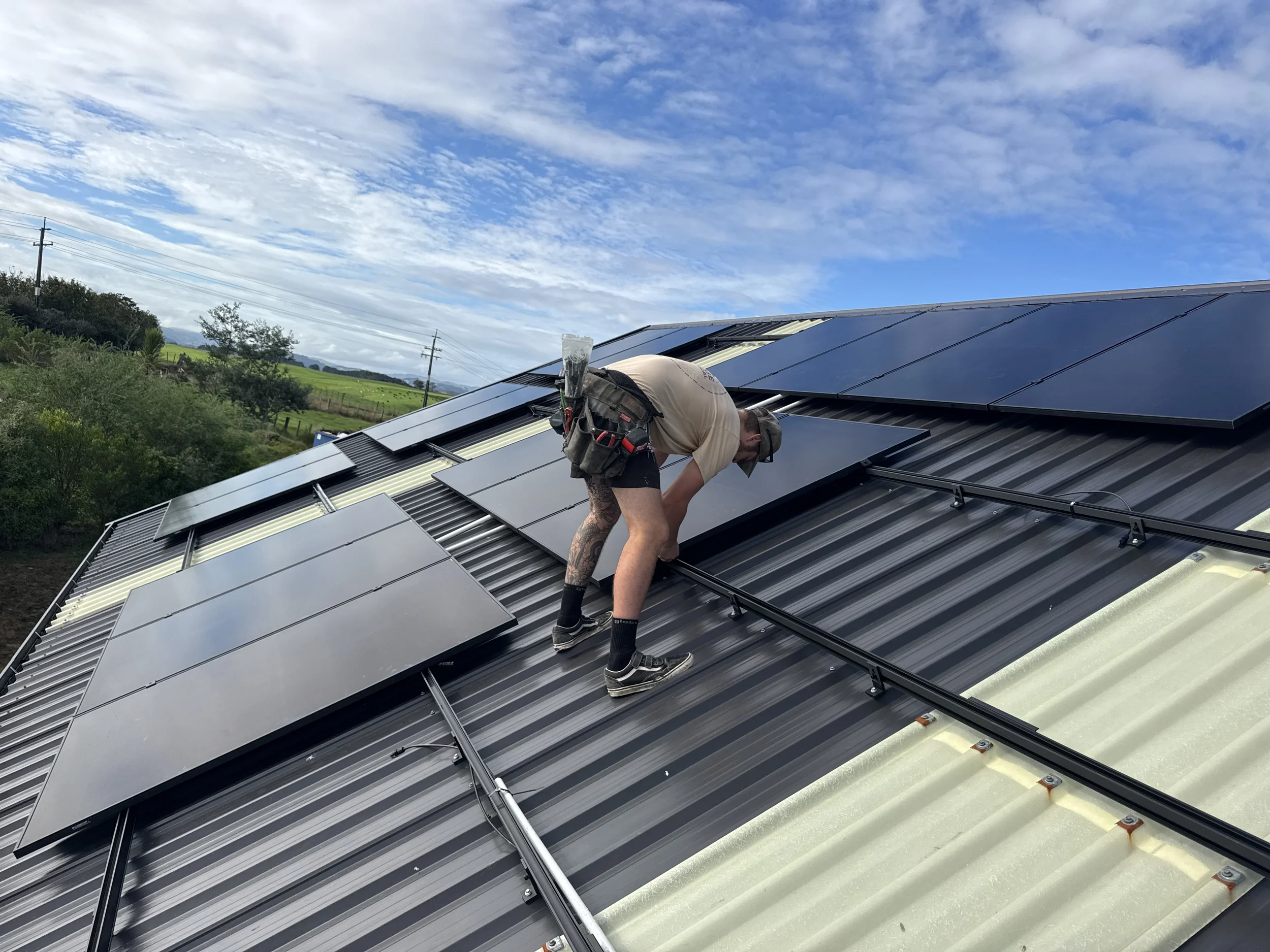 A person installing solar panels on a metal roof during daytime with a partly cloudy sky.