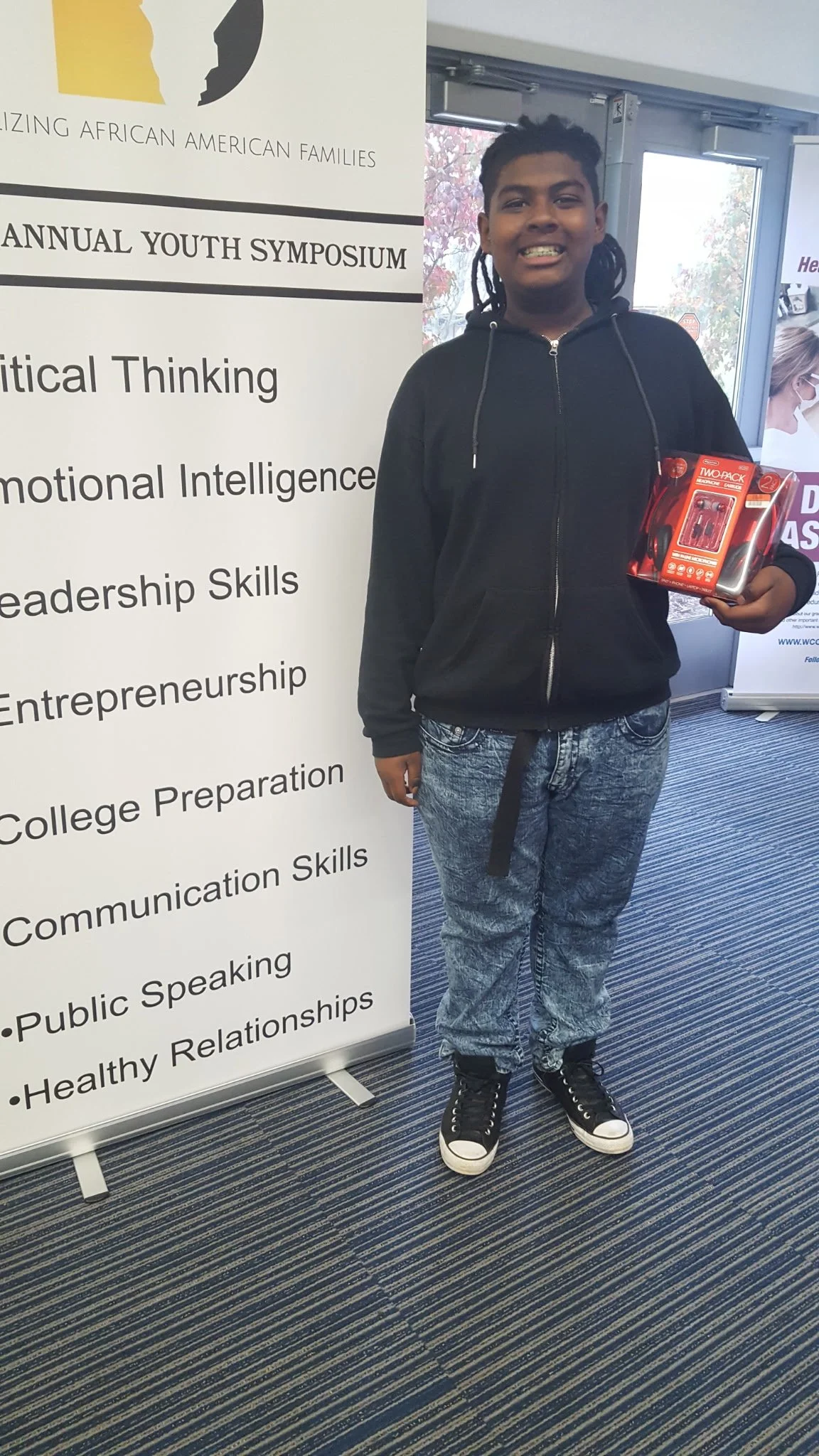 A smiling young man with dreadlocks, wearing a black hoodie, jeans, and high-top sneakers, standing indoors next to a banner promoting an African American families youth symposium.