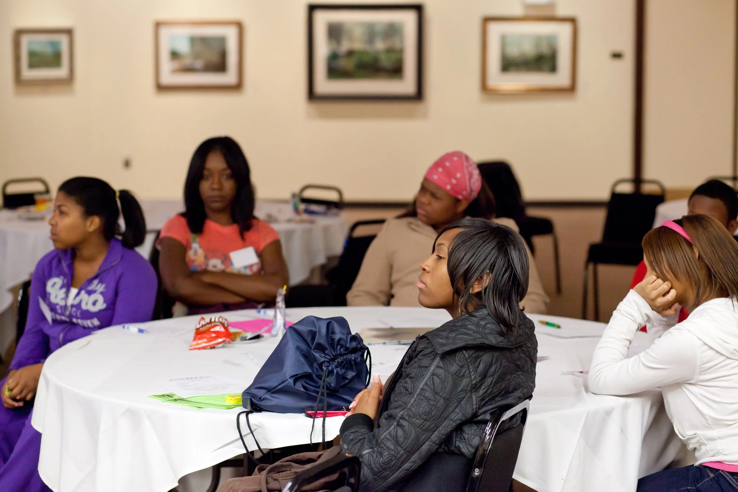 Group of women sitting at round tables during a gathering or meeting in a conference room, some appear to be listening attentively.