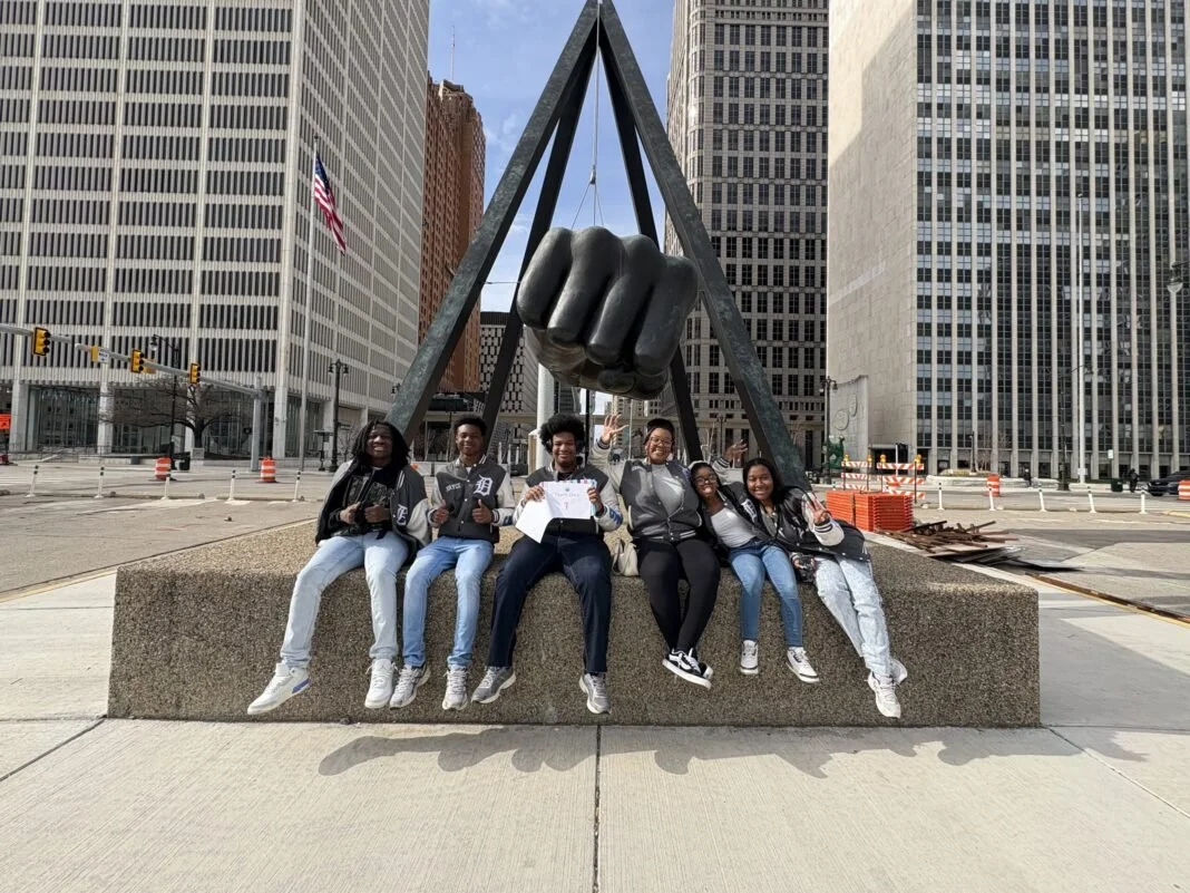 Group of six young people sitting on a large concrete platform in front of a sculpture of a fist lifting weights, surrounded by tall city buildings.