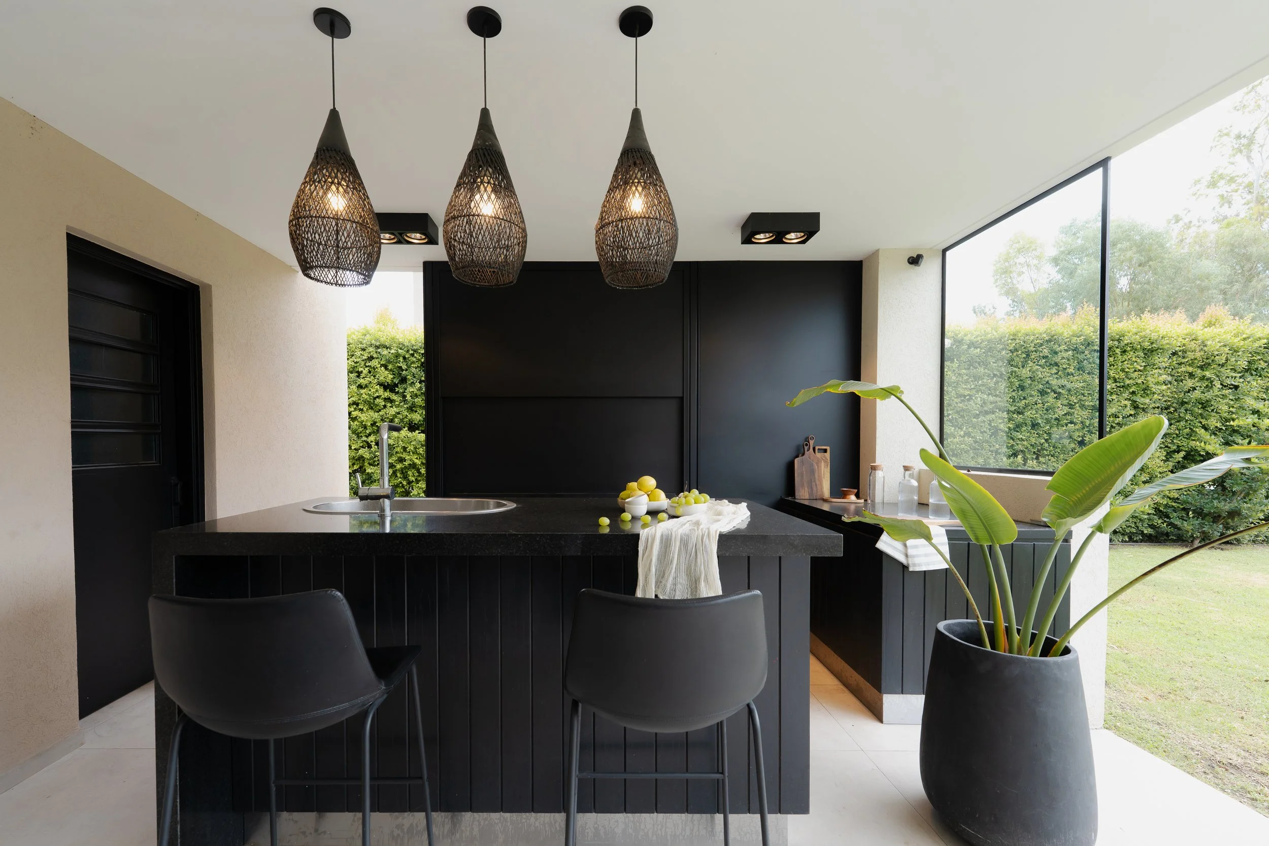 Cocina moderna con isla negra, lámparas colgantes de mimbre y gran ventana con vista a un jardín con arbustos verdes.