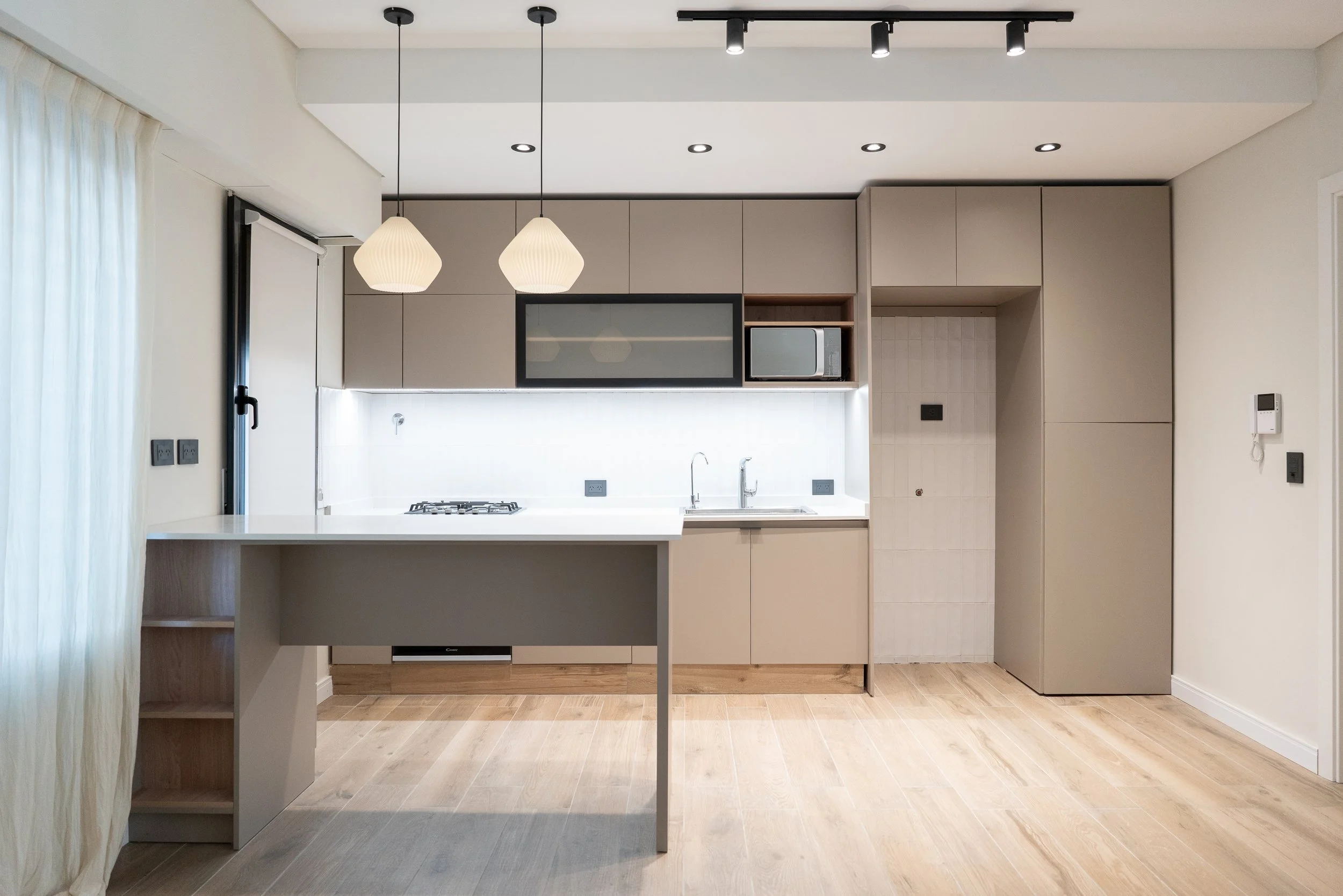 Cocina moderna con gabinetes beige, estantería de microondas, fregadero, estufa y placas eléctricas, con lámparas colgantes y piso de madera clara.