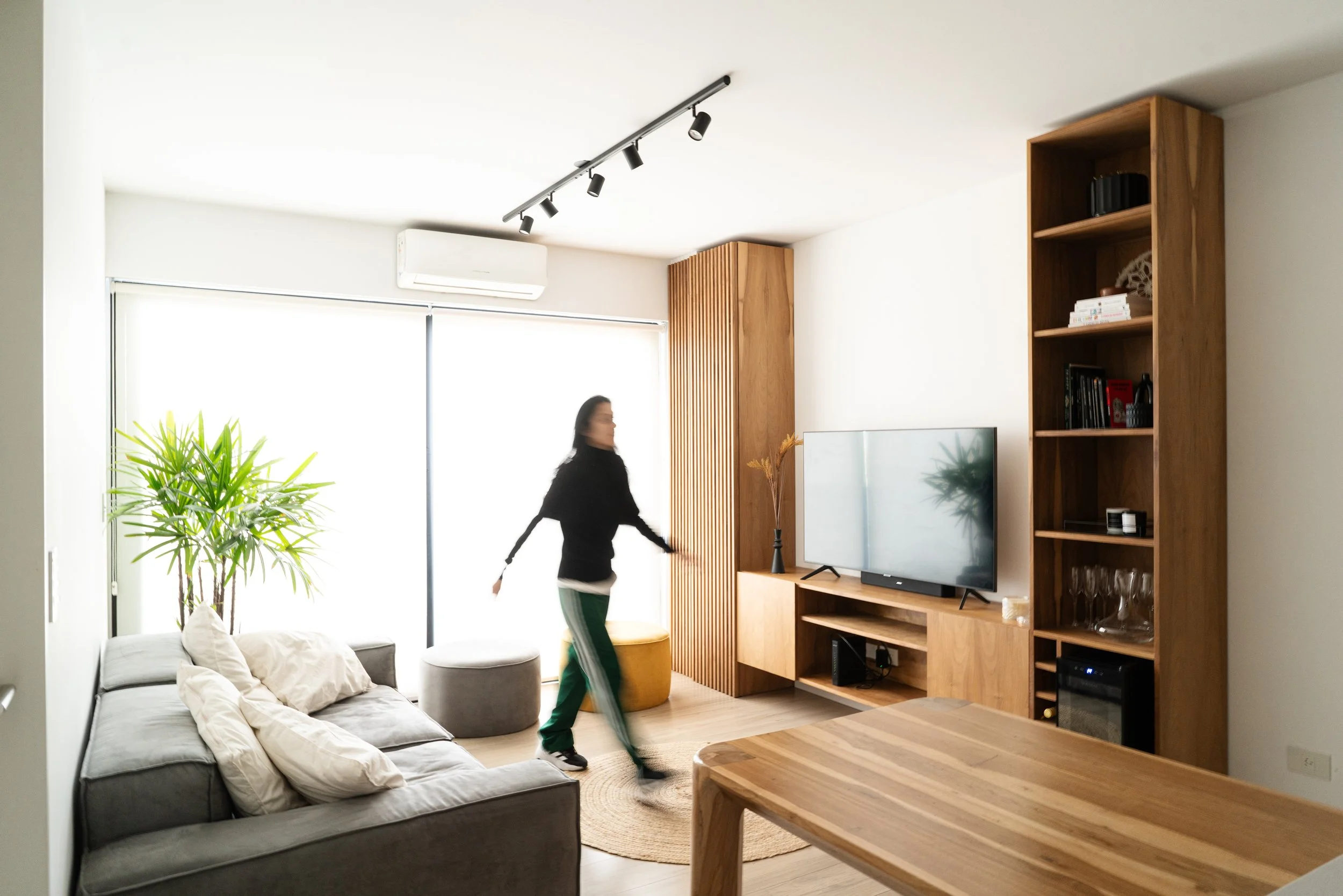 Sala moderna con sofá gris, plantas verdes, televisión en mueble de madera, estanterías y ventanas con cortinas blancas, mujer caminando