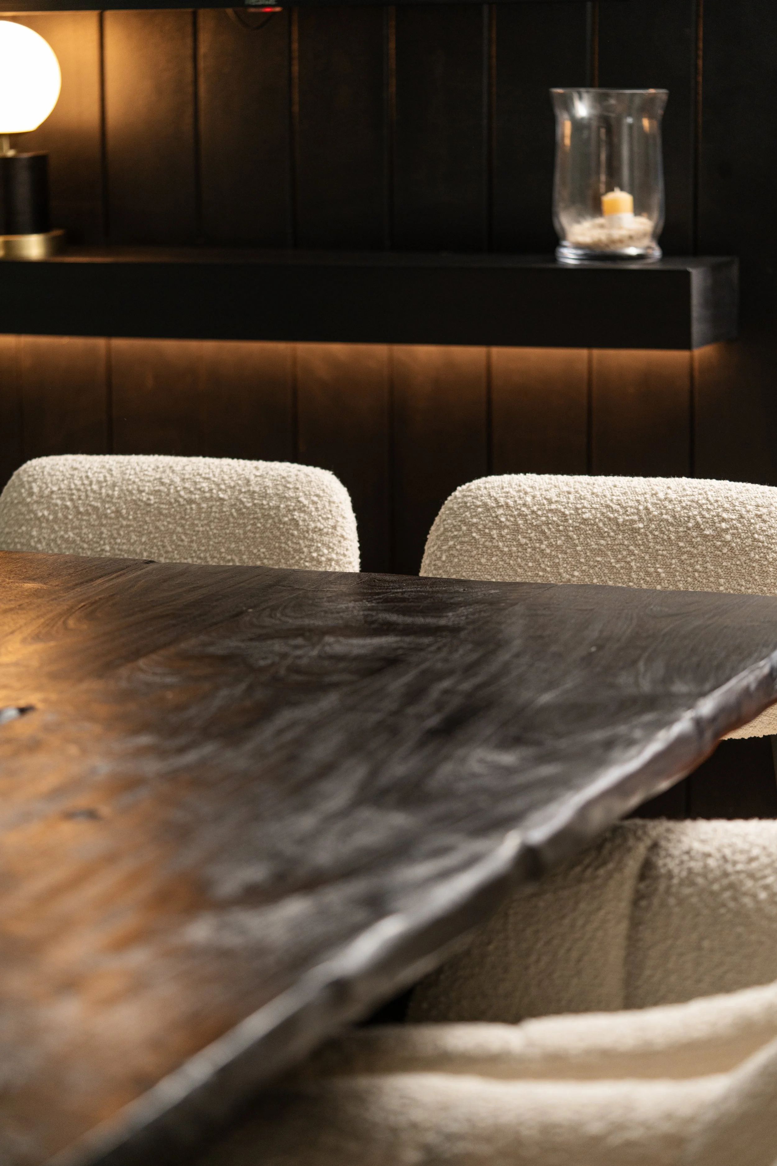 Vista de una mesa de madera oscura y sillas con tapizado beige en un ambiente interior con pared de madera y decoración con luz cálida y un jarrón con vela.