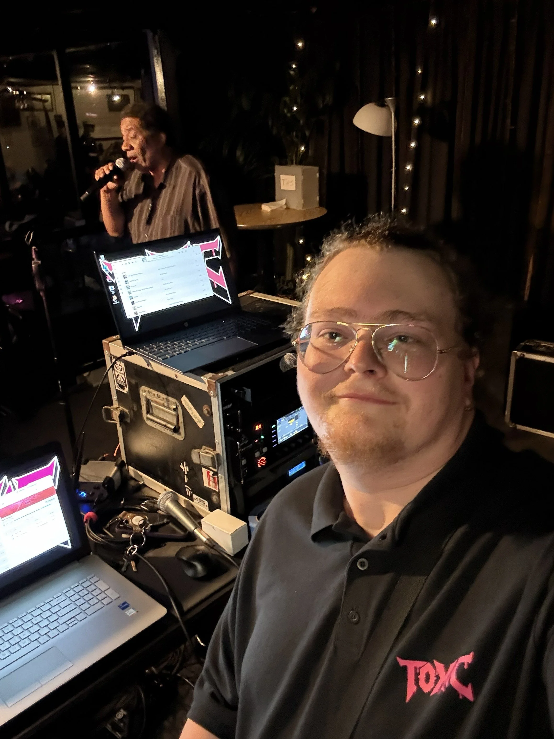 A person with glasses taking a selfie at a control station with computers and a woman singing into a microphone in the background.