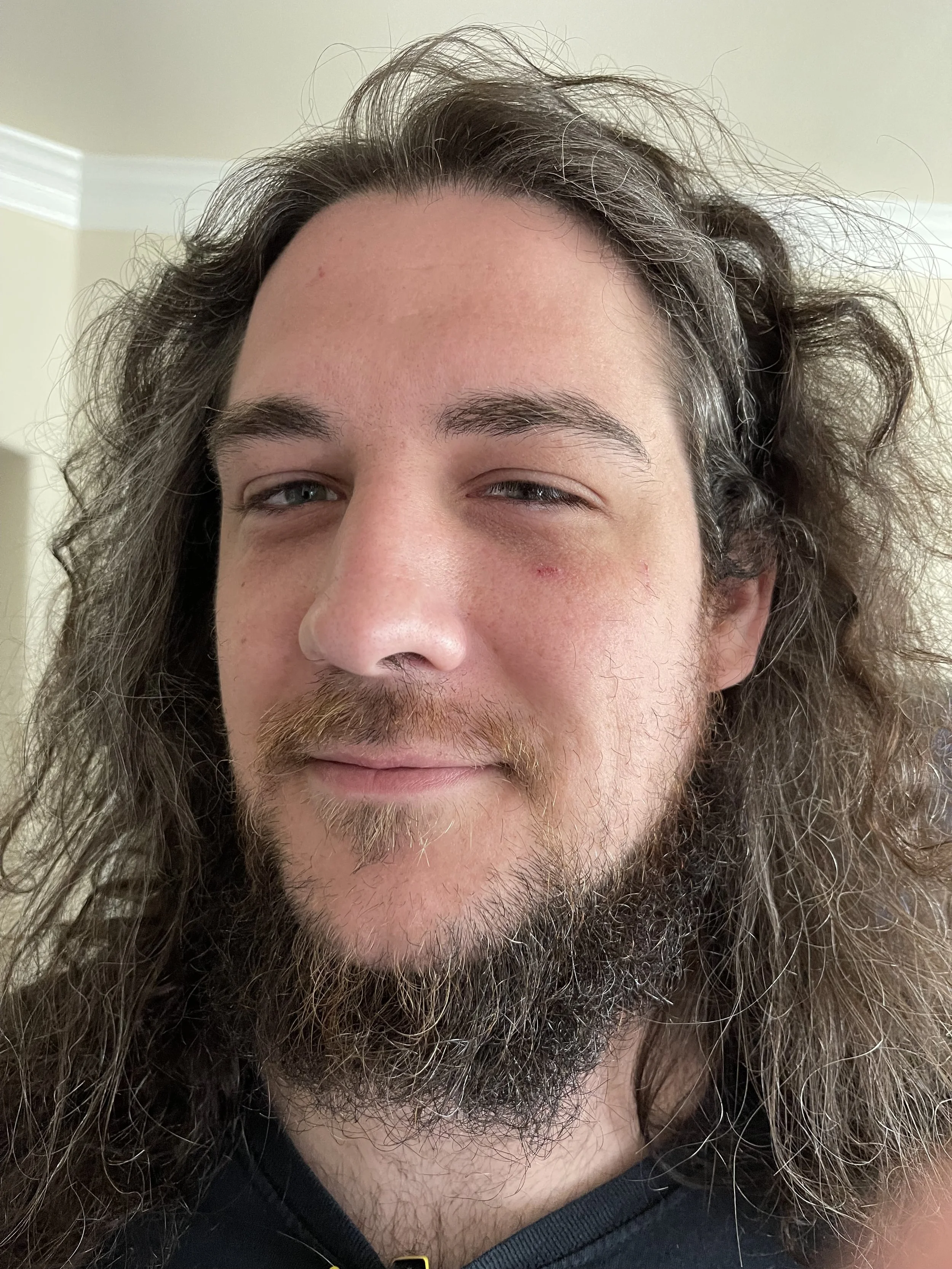 Close-up of a person with long, curly hair, partially winking, with some facial hair and a beard, wearing a dark top.