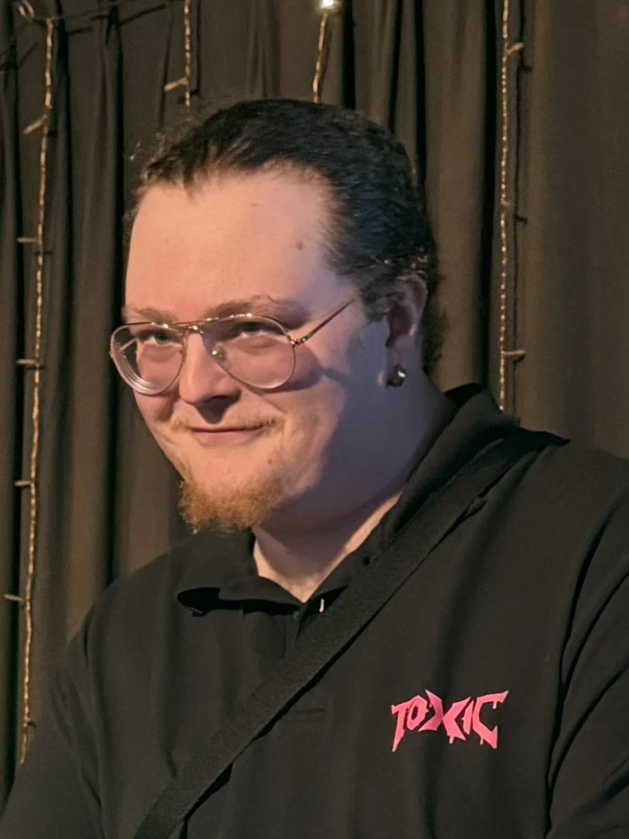 A person with glasses, dark slicked-back hair, a beard, and earrings, smiling slightly against a dark curtain background wearing a black shirt with a pink logo.