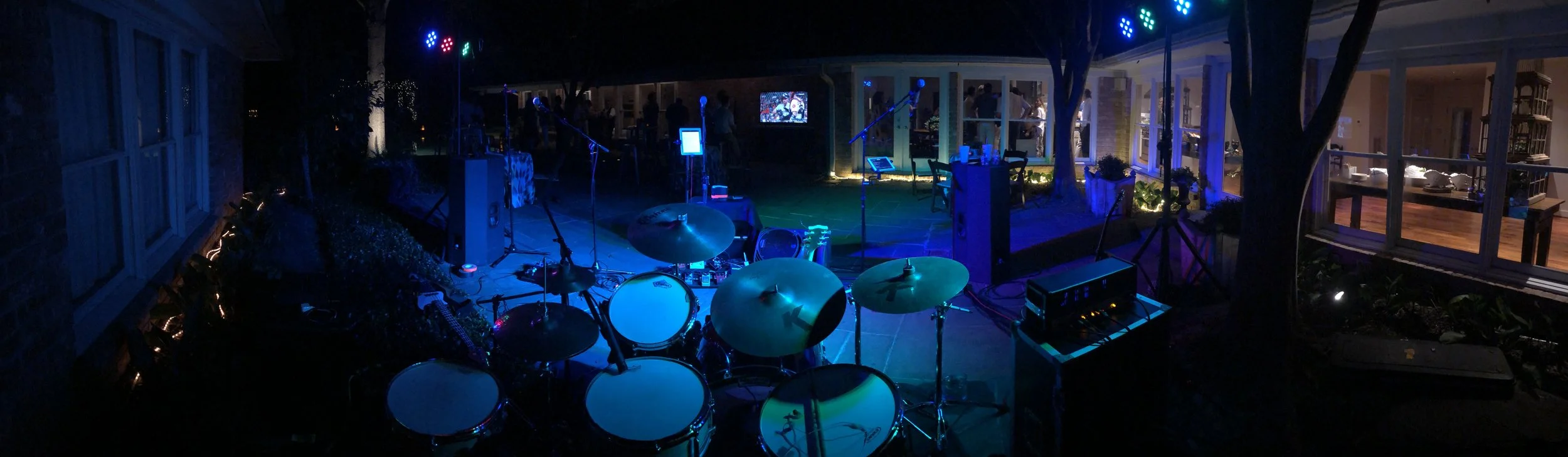 Outdoor nighttime scene of a stage set with musical instruments, lighting, and microphones, with a group of people gathered around inside a glass-enclosed room.
