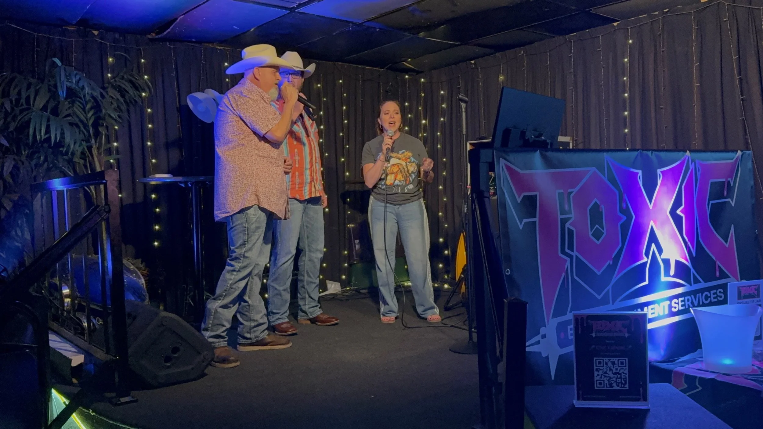 Three people on stage at a performance venue, with the man on the left wearing a cowboy hat and light-colored shirt, holding a microphone. The woman on the right also holding a microphone and wearing a graphic t-shirt. The background features dark cu