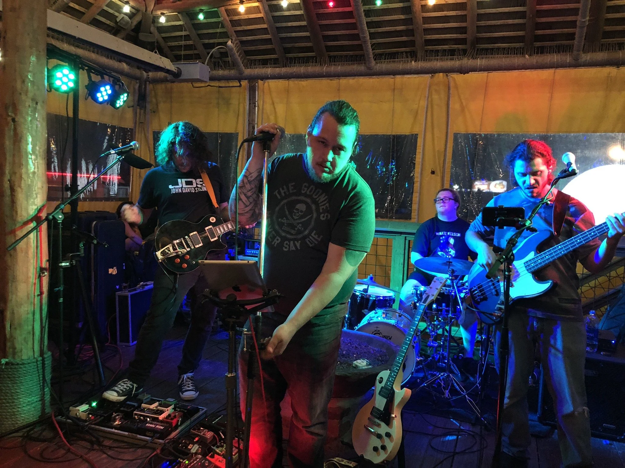 A band performing live on stage with instruments including electric guitar, bass, and drums, under colorful stage lighting in a cozy venue.