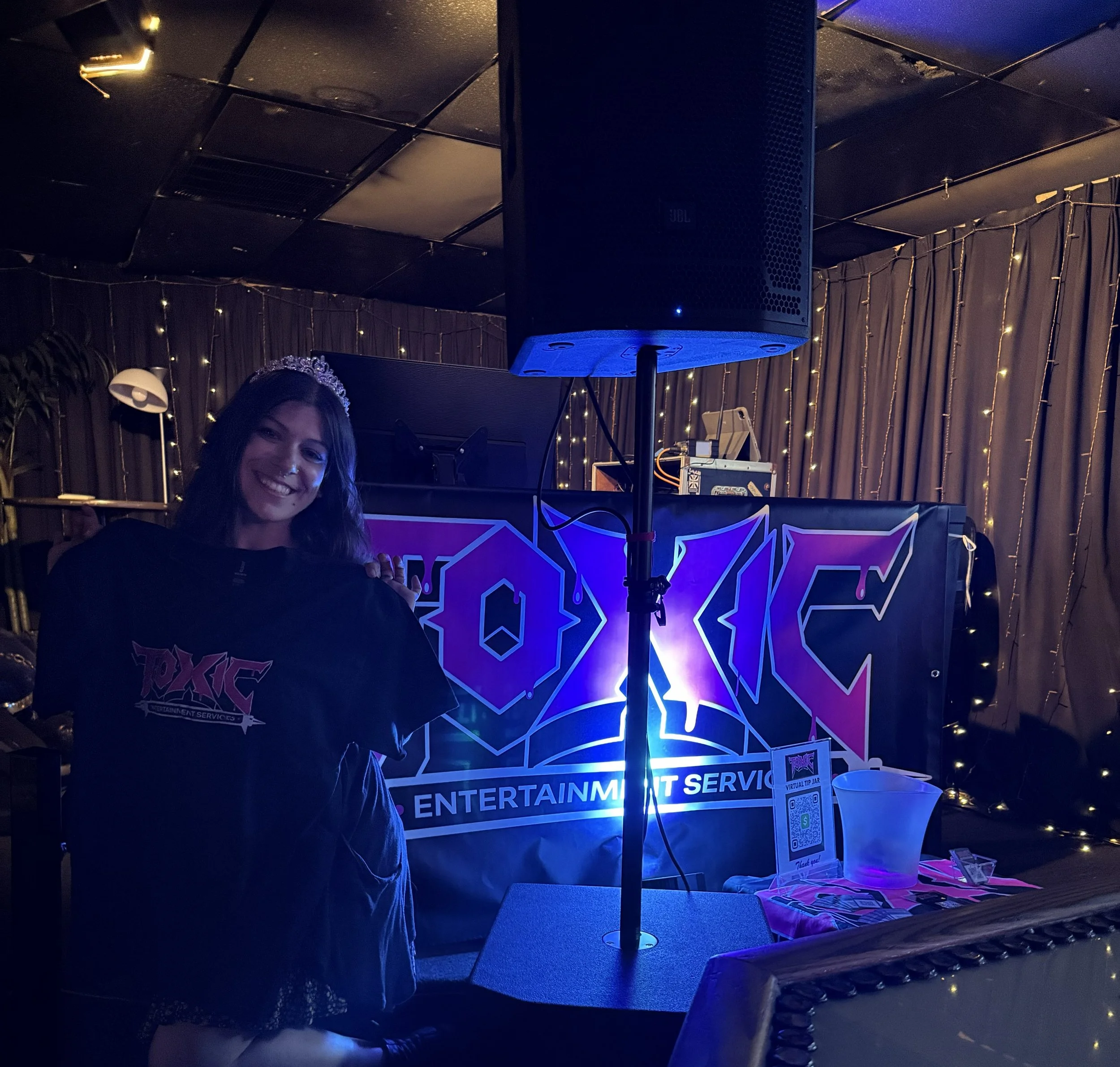 A smiling young woman wearing a tiara standing near a DJ booth at a party or event with decorative string lights and a sign that reads 'ENTERTAINMENT'.