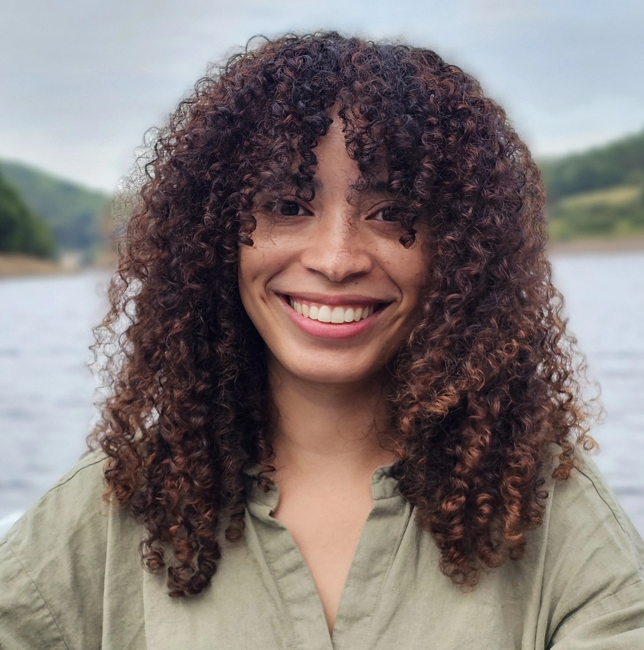 Kiersten Formoso, a smiling woman with curly brown hair outdoors with a lake and green hills in the background.