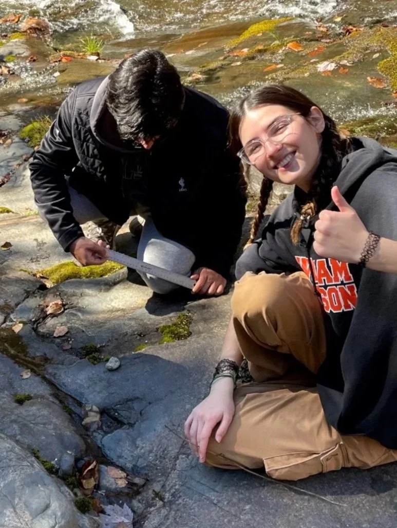 Two people outdoors atop dirt, one sitting with a hooded sweatshirt giving a thumbs-up and smiling at the camera and the other kneeling with a tool.