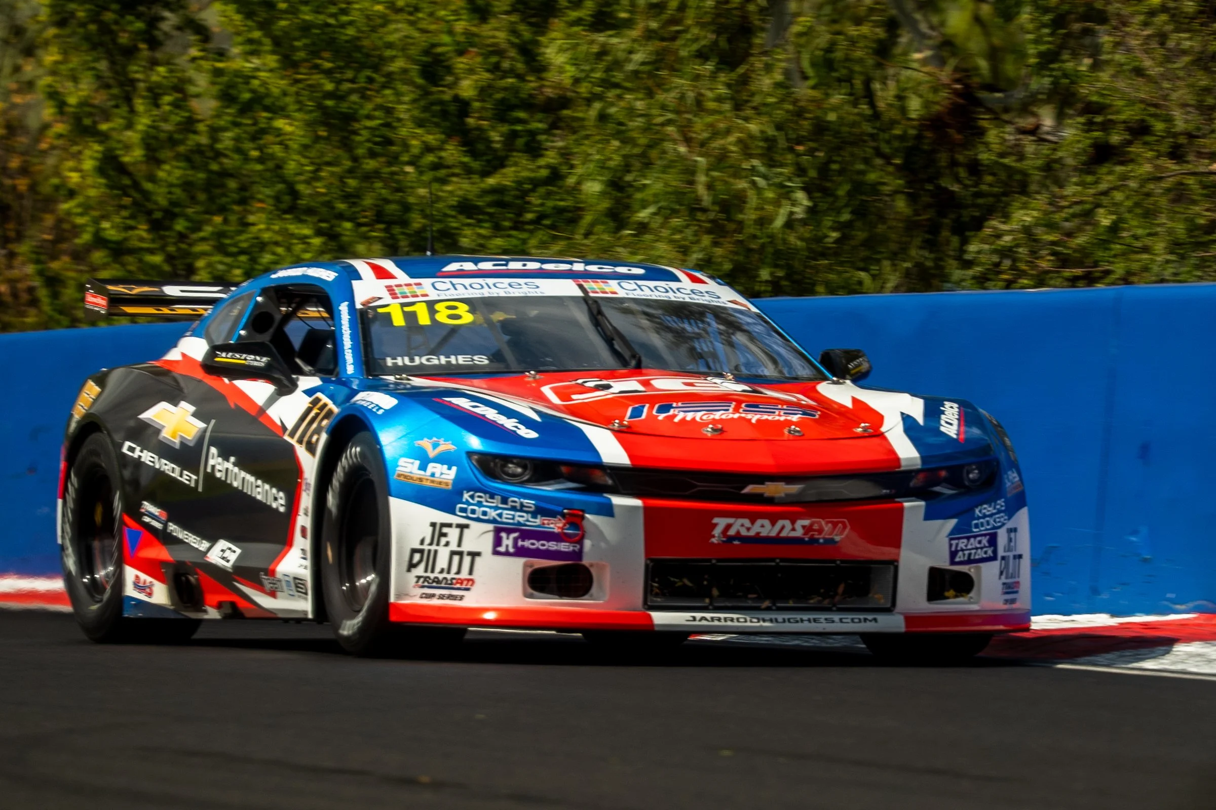 Red, white and blue Chevrolet race car number 118 navigating a curve on the racetrack, with a blue barrier and green vegetation in the background