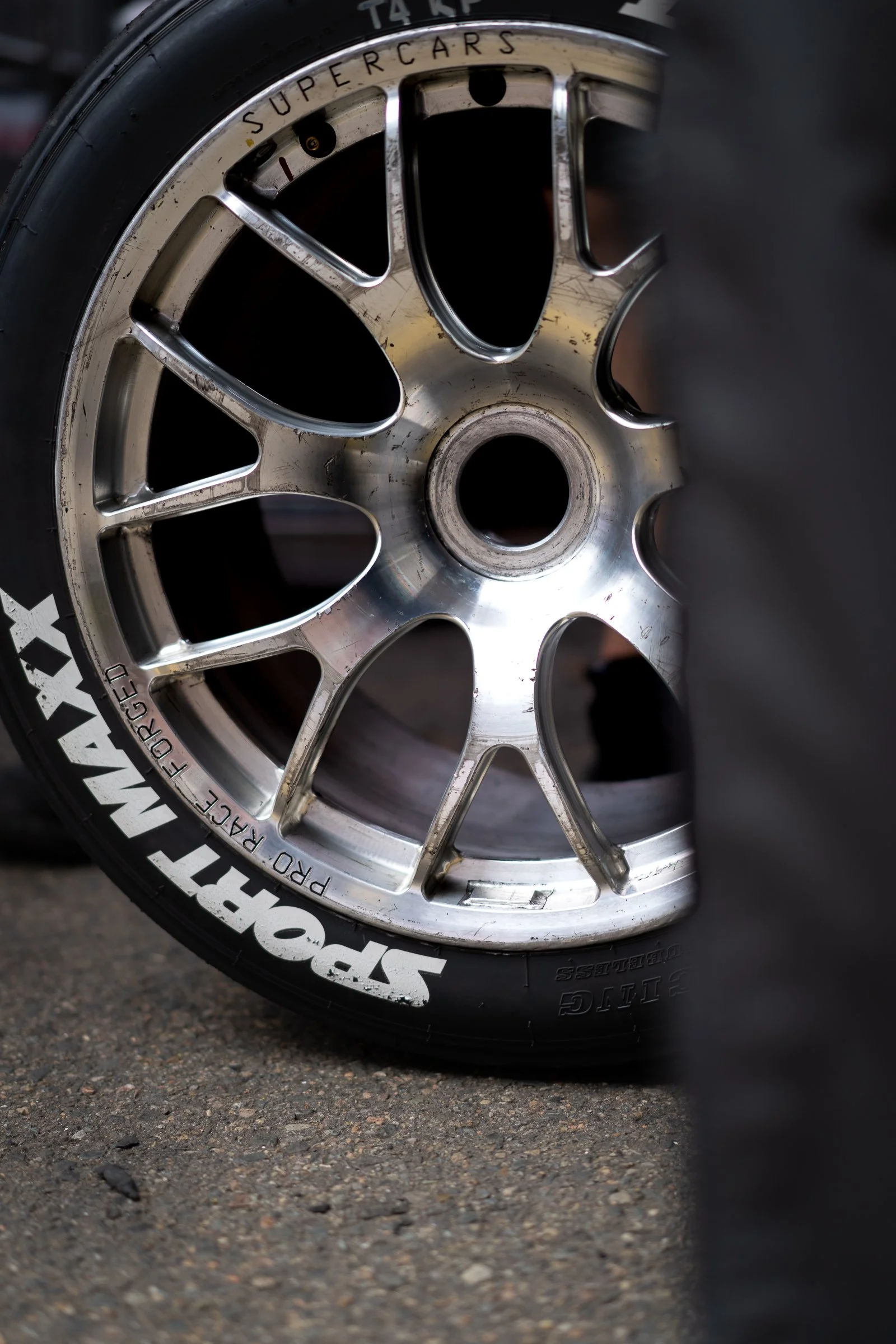 lose-up of a worn multi‑spoke racing wheel with “SPORT MAXX” lettering on the tyre and “PRO RACE FORGED” and “SUPERCARS” markings visible on the rim, resting on asphalt