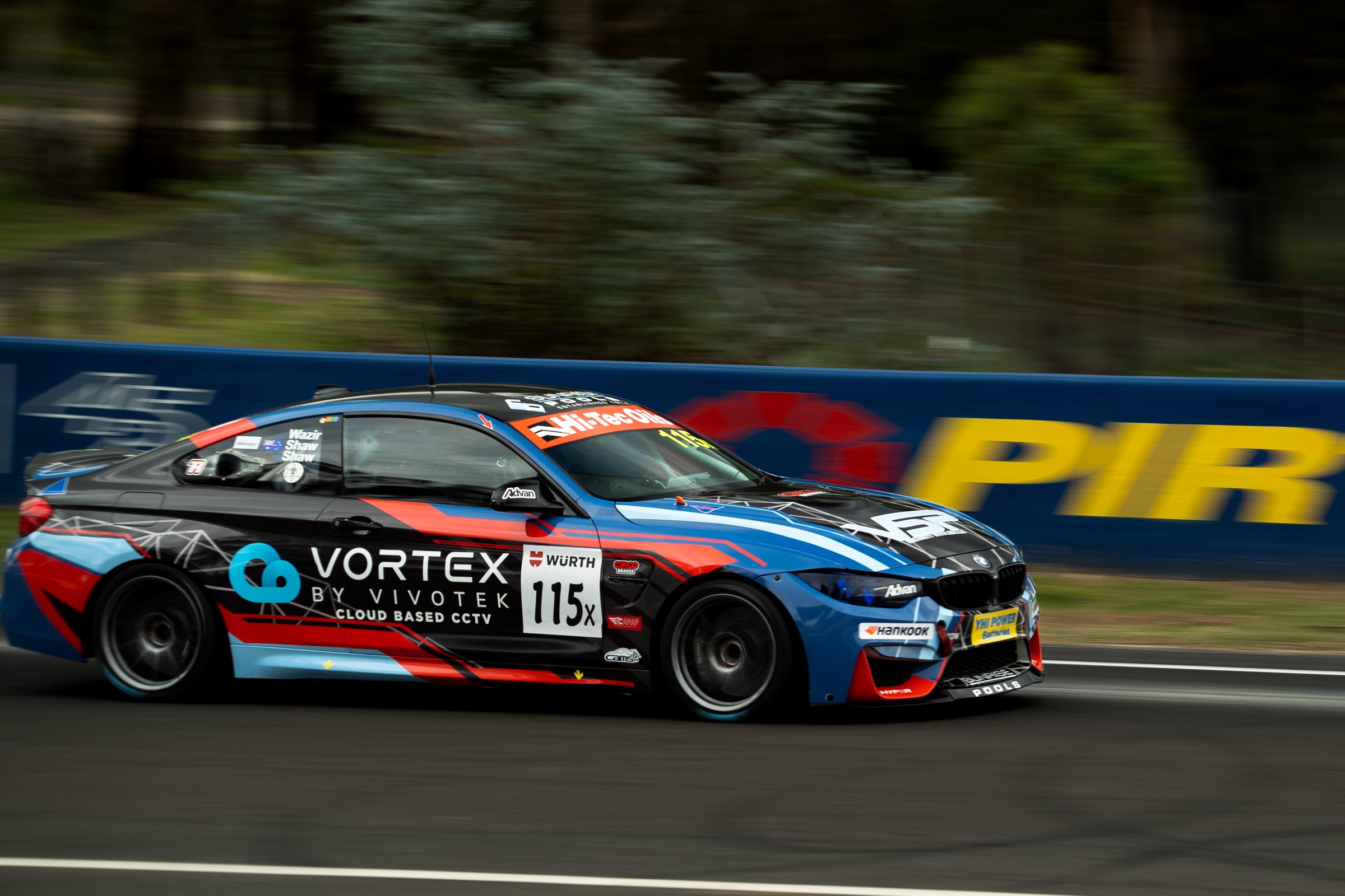 Side view of the number 115 BMW race car on track, passing a barrier with Pirtek branding, showing its blue, black and red livery and sponsor logos