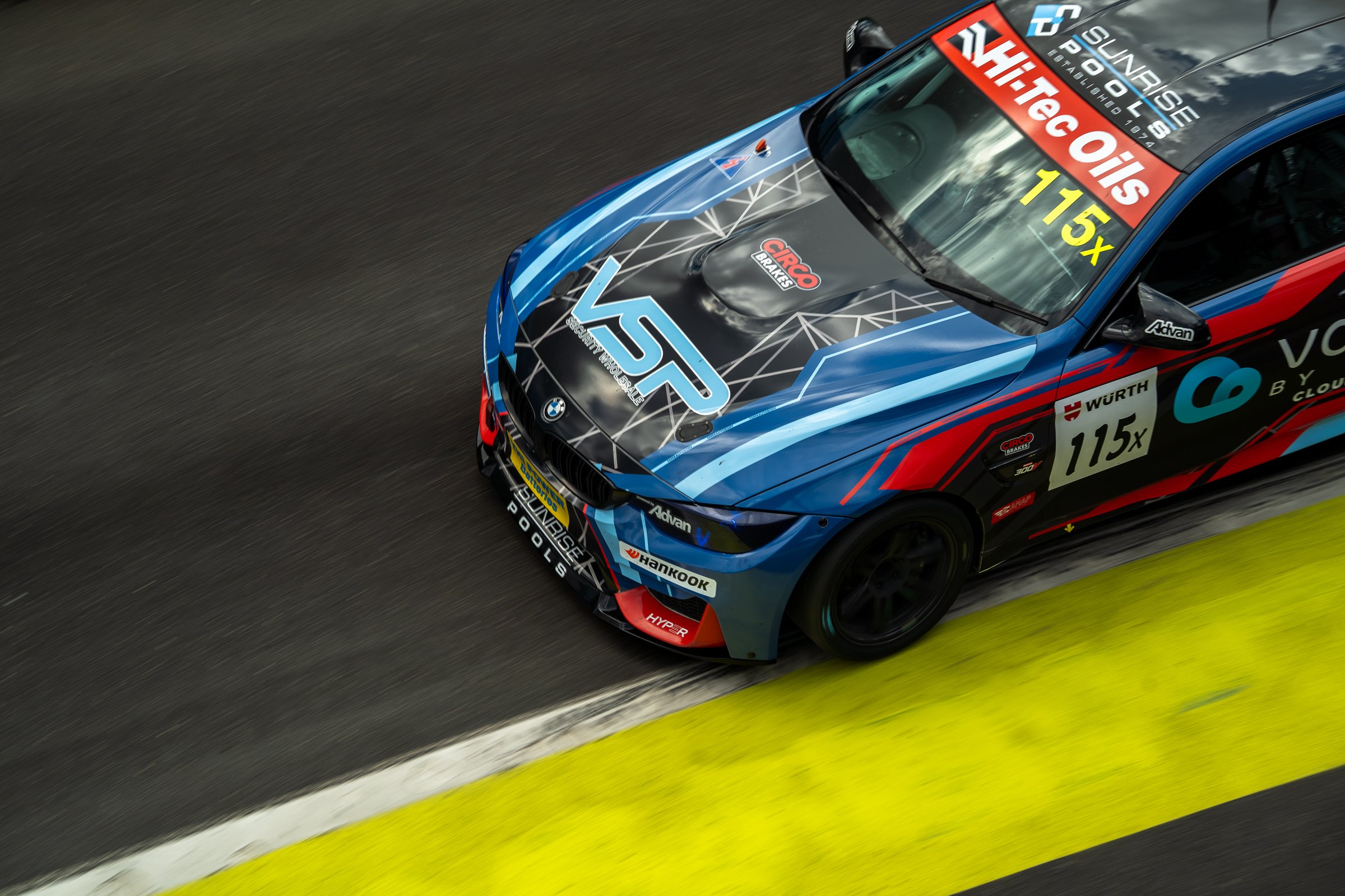 Overhead view of the number 115 BMW race car on track, running close to the yellow line with its blue, black and red livery and sponsor logos visible
