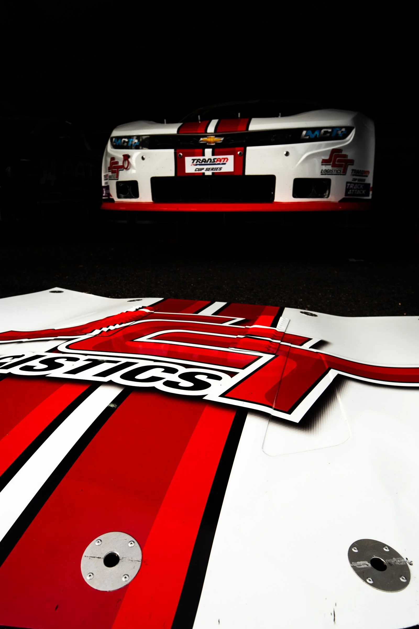 Close‑up of a race car bonnet featuring a large SCT Logistics logo with red, black and white stripes, with the front of the car visible in a dark garage