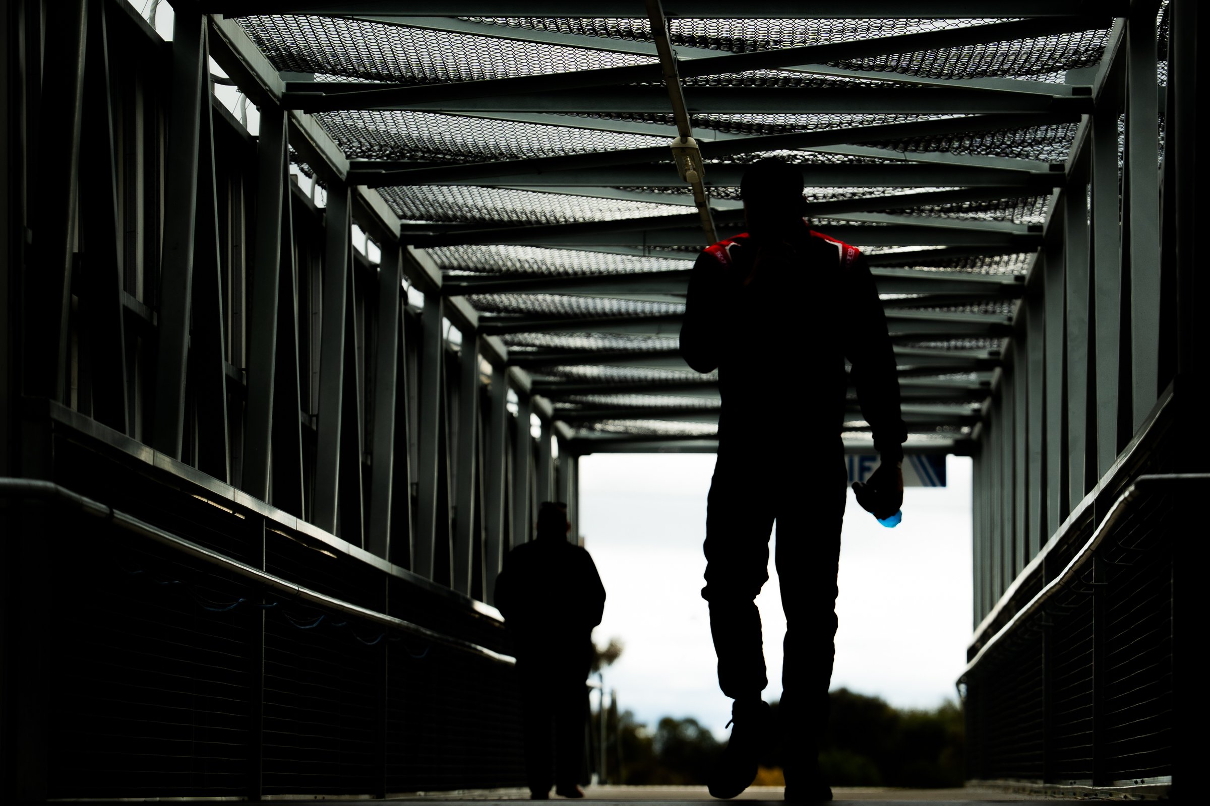 Silhouetted person walking toward the camera through a metal‑framed covered walkway at the Bathurst 6 Hour