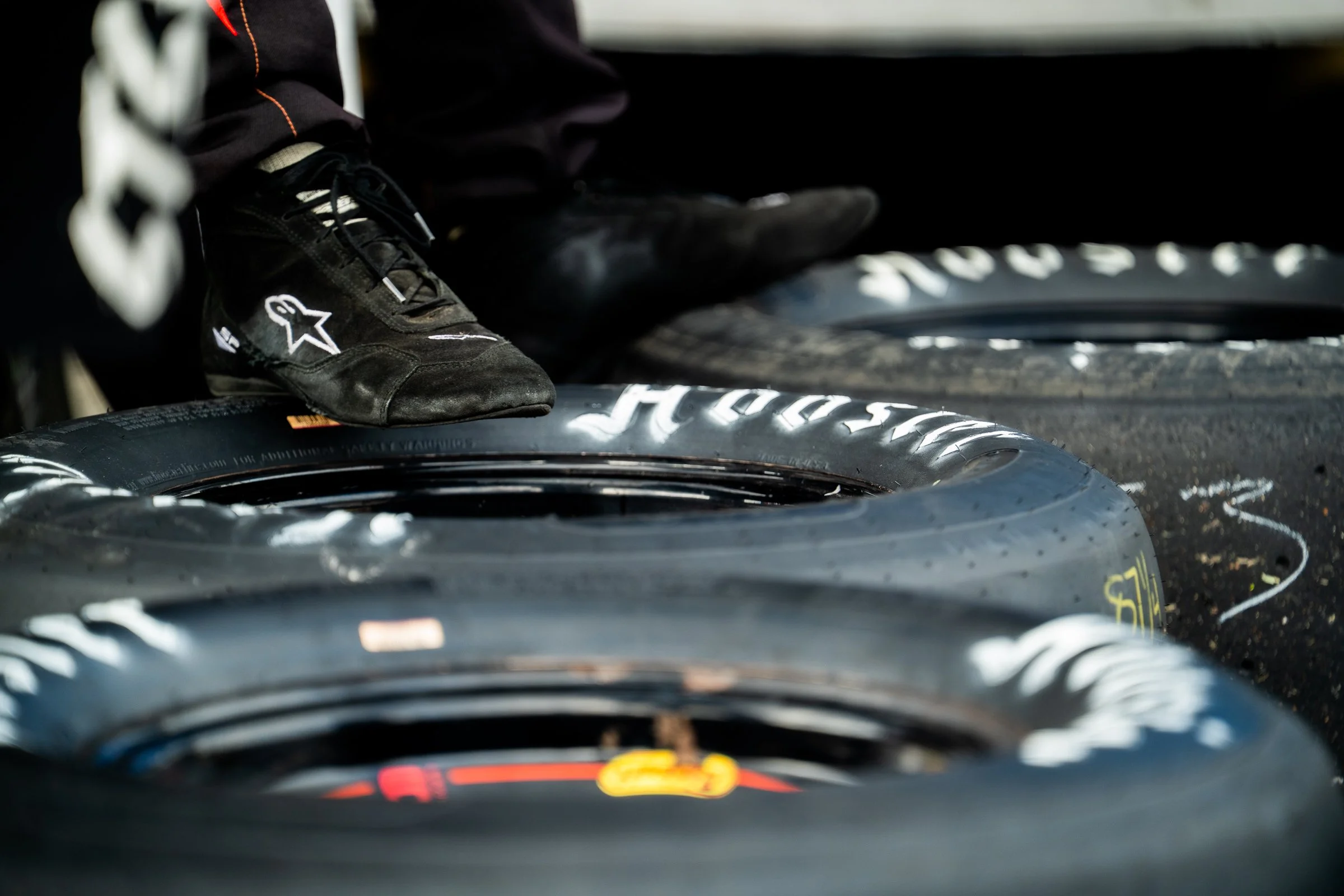 Close-up of a race car driver's foot on a race car tire, wearing a black racing shoe with a white logo, next to a row of worn racing tires