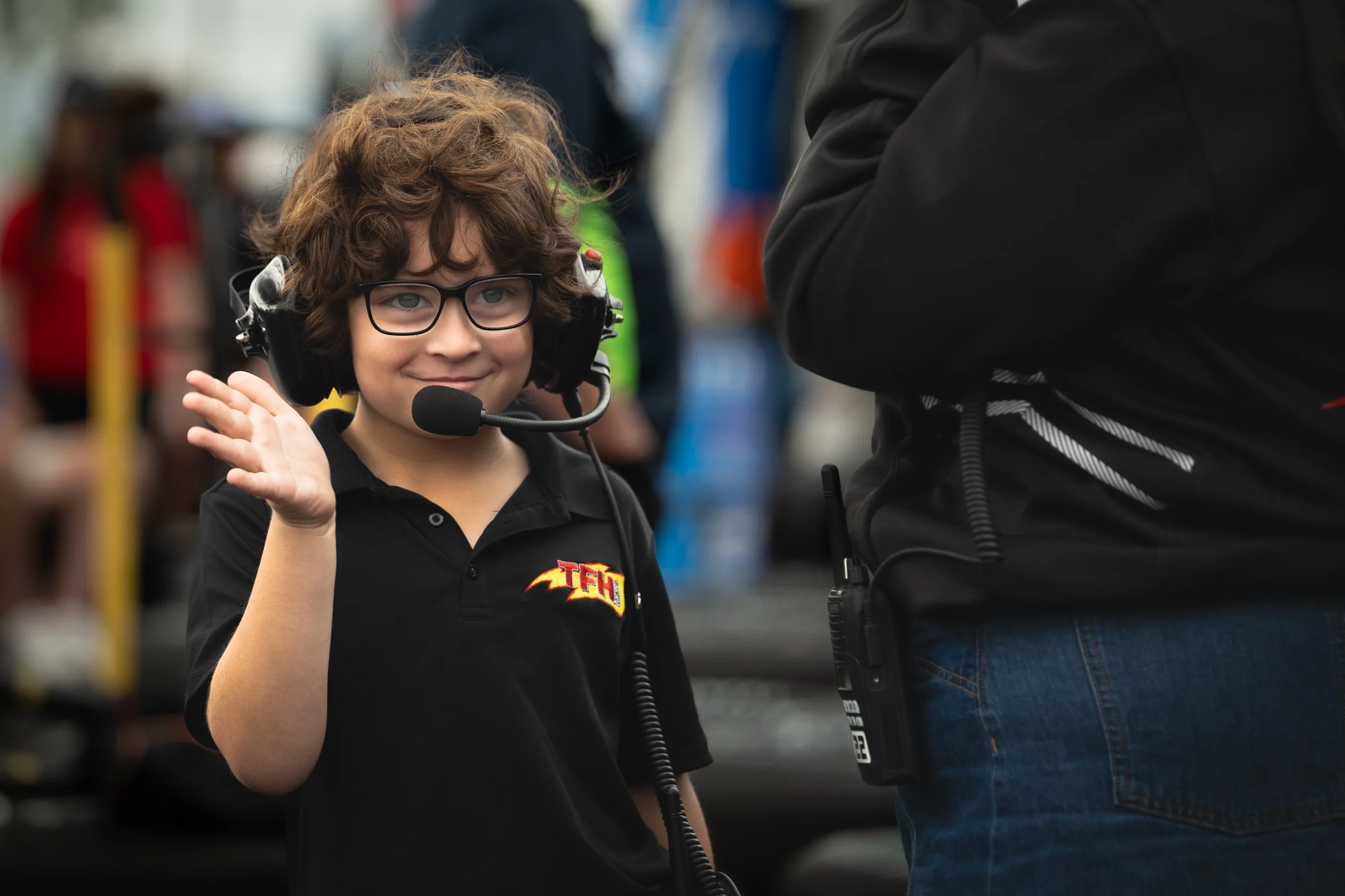 Young boy with curly hair wearing glasses and a headset waving and smiling beside a person in black clothing with equipment on their belt