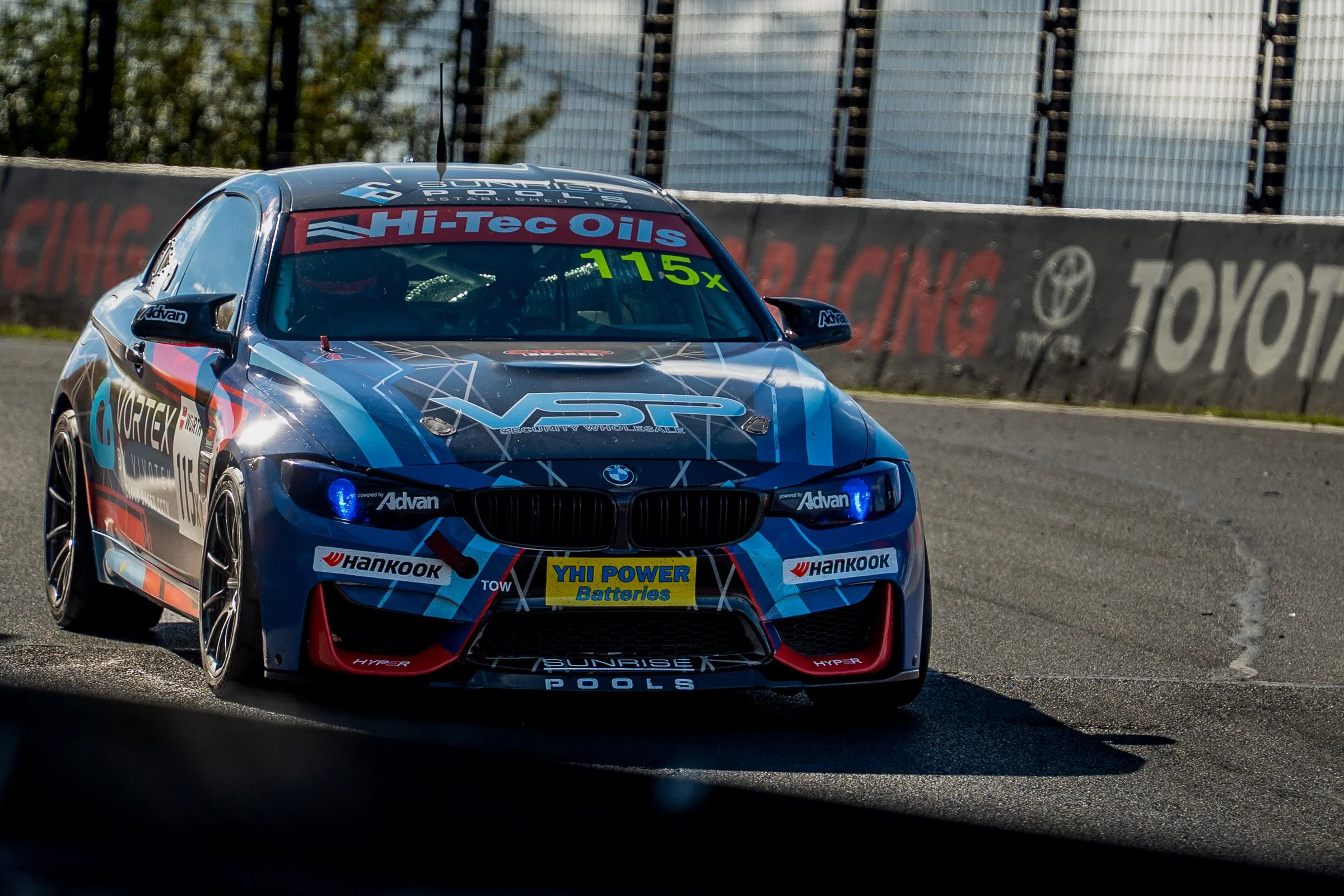 Blue BMW race car with Hankook and Hi‑Tec Oils branding navigating a turn on the racetrack during daytime