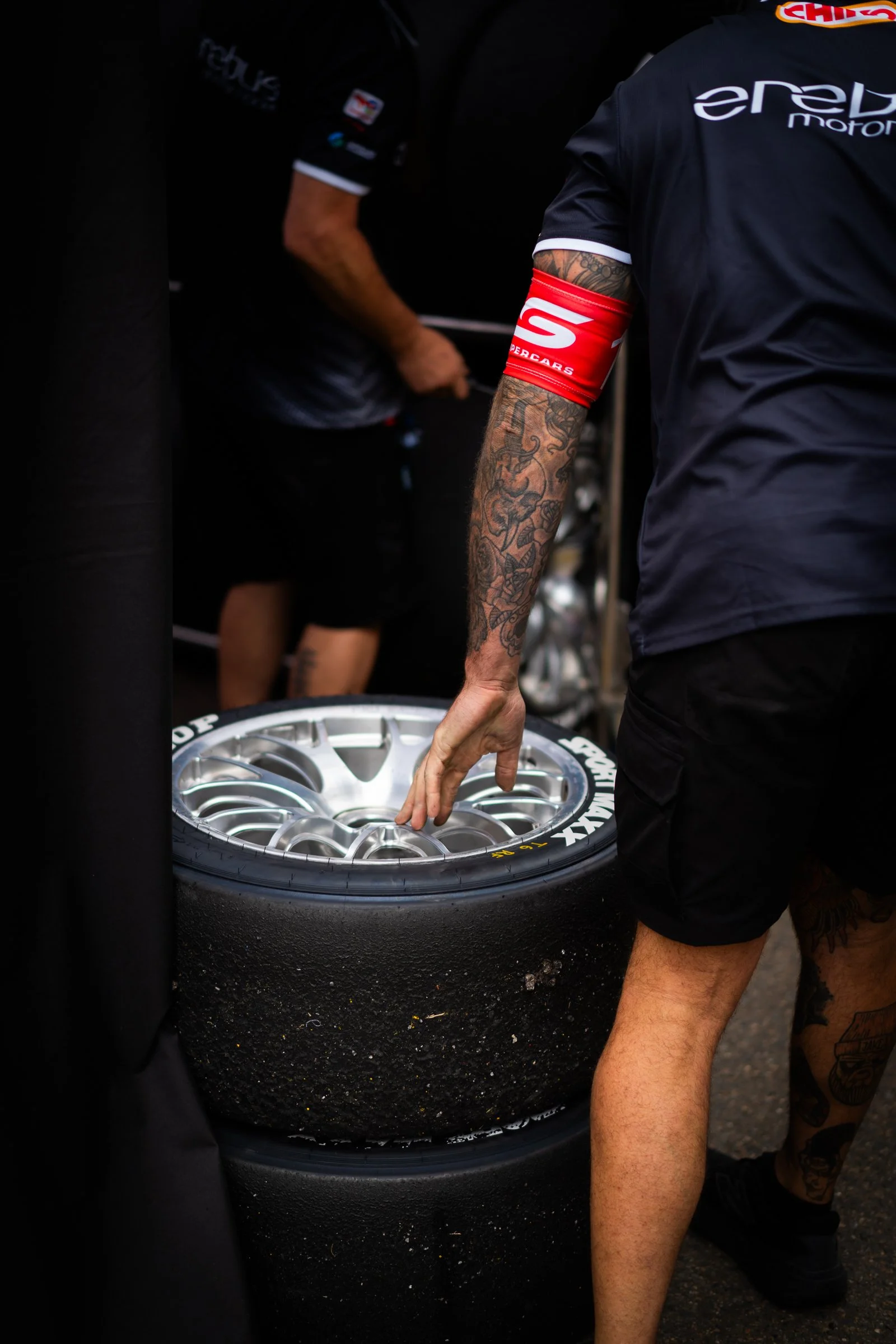 Erebus Motorsport crew member handling stacked Dunlop Sport Maxx racing tyres in the garage area, with another team member in the background