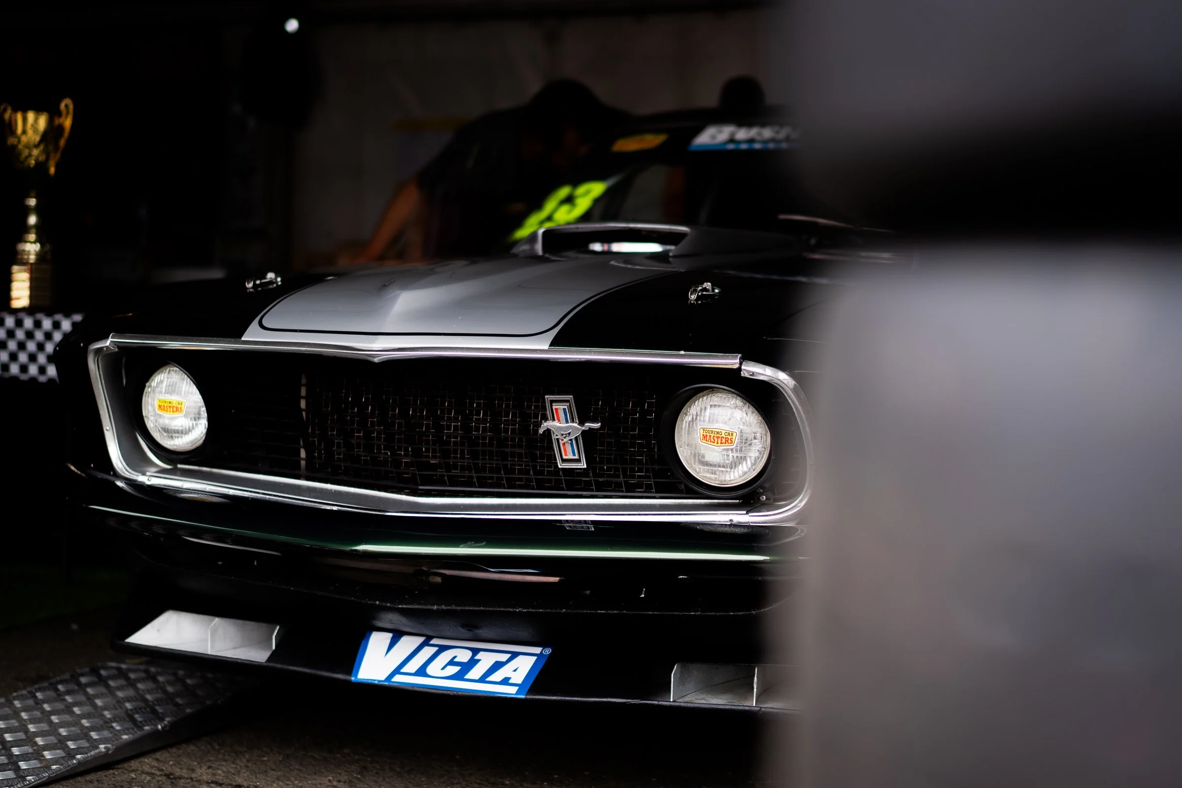 Close‑up of a black vintage Ford Mustang race car in a dark garage, partially obscured by stacked tyres, with a VICTA sign on the bumper and a trophy on a nearby table