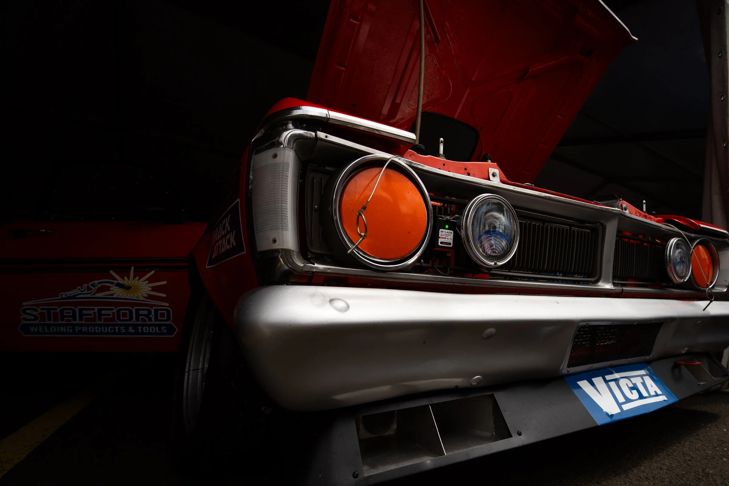 Close‑up of an orange vintage race car with the bonnet open showing the headlights and radiator grille under a tent
