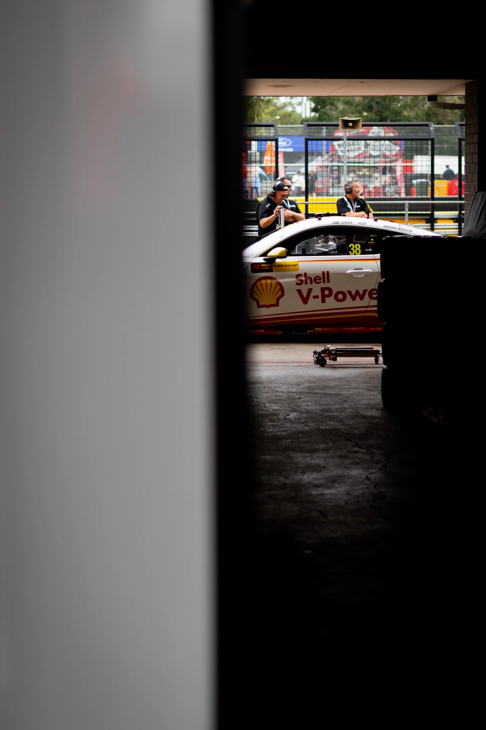 Two men wearing headphones near a Shell V‑Power–branded race car, viewed through a garage with silhouetted equipment in the foreground