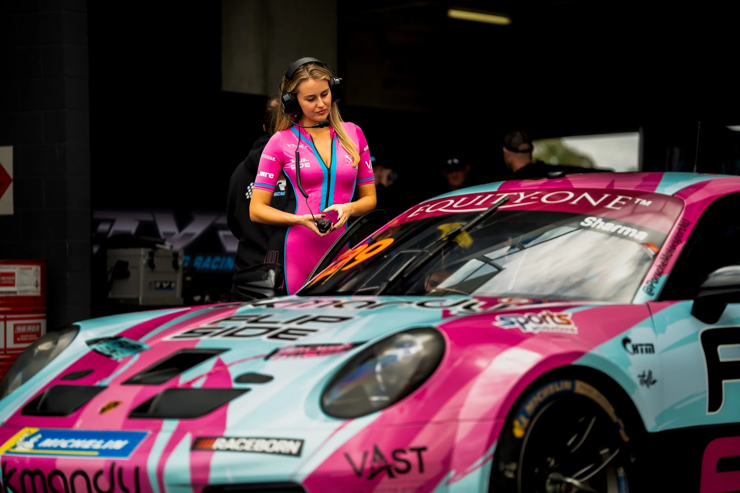 Promotional model in a pink and blue bodysuit wearing a headset standing near a pink and blue race car in front of a garage